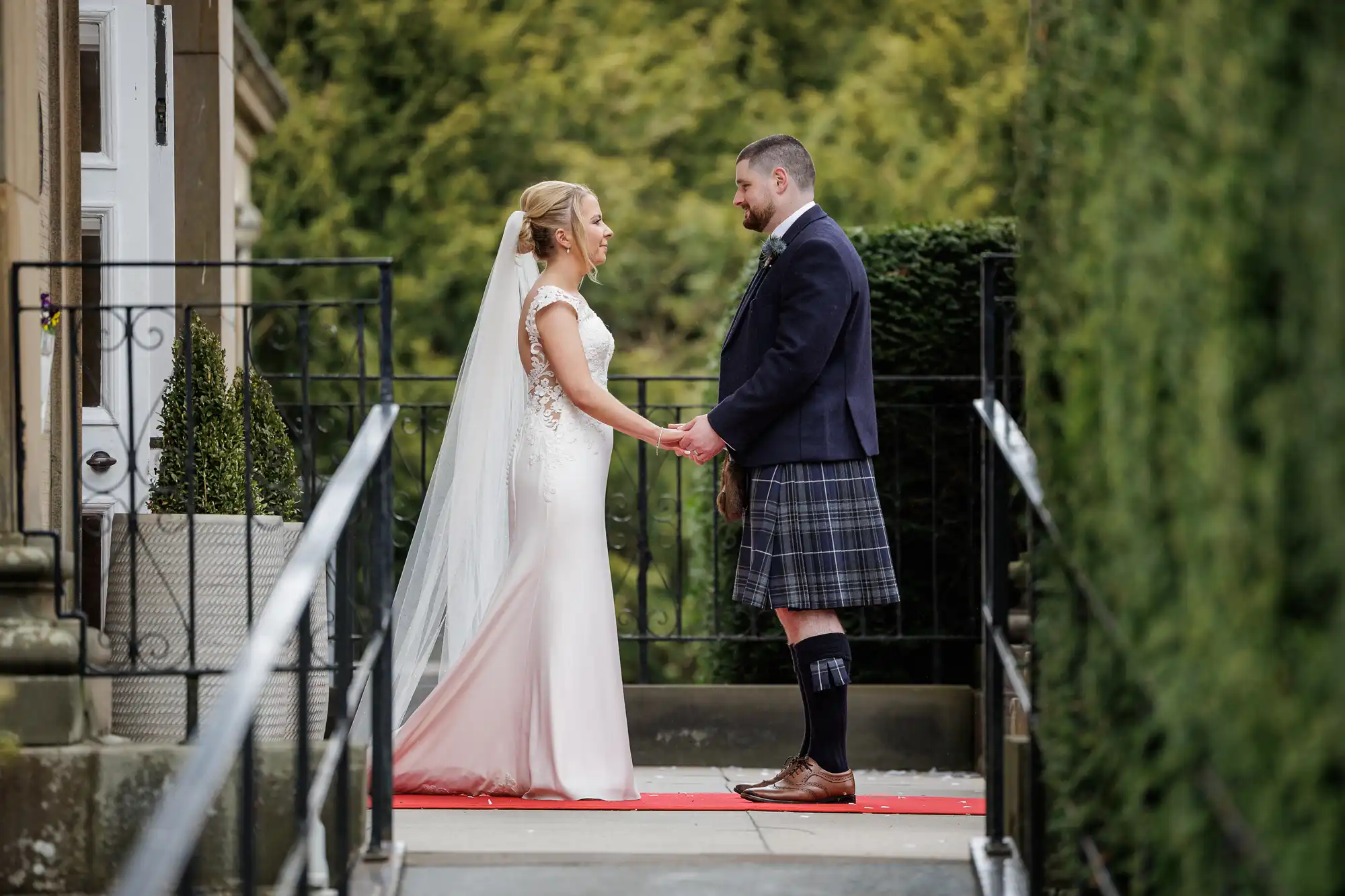 A bride in a white dress and veil holds hands with a groom in a dark jacket, kilt, and knee-high socks. They face each other on a red carpet, outdoors, surrounded by greenery and wrought iron railings, sharing an intimate moment.