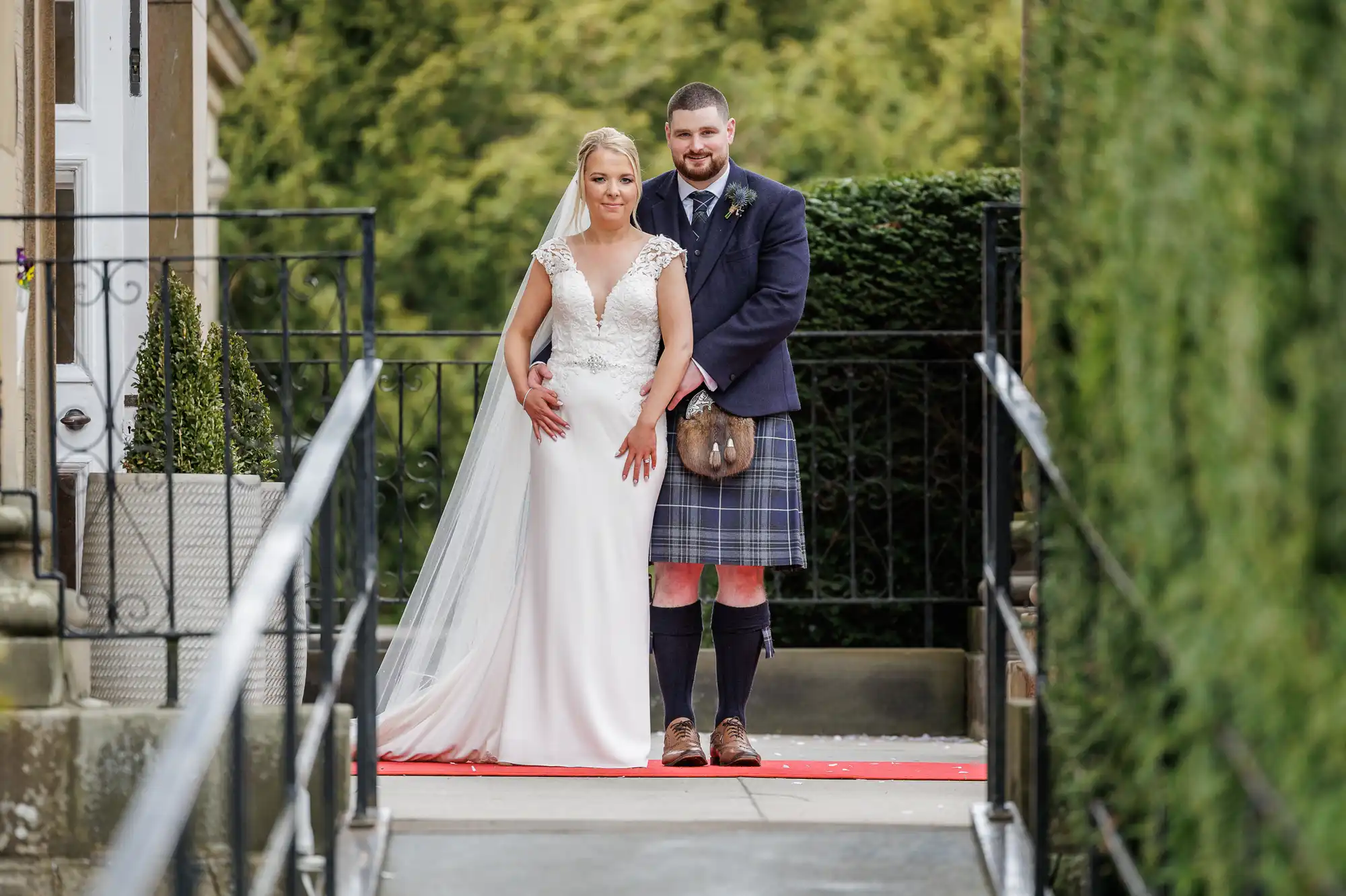 A bride in a white dress and veil stands in front of a groom wearing a dark jacket and traditional Scottish kilt. They pose together on a red carpet outdoors, with greenery and a stone building visible in the background.