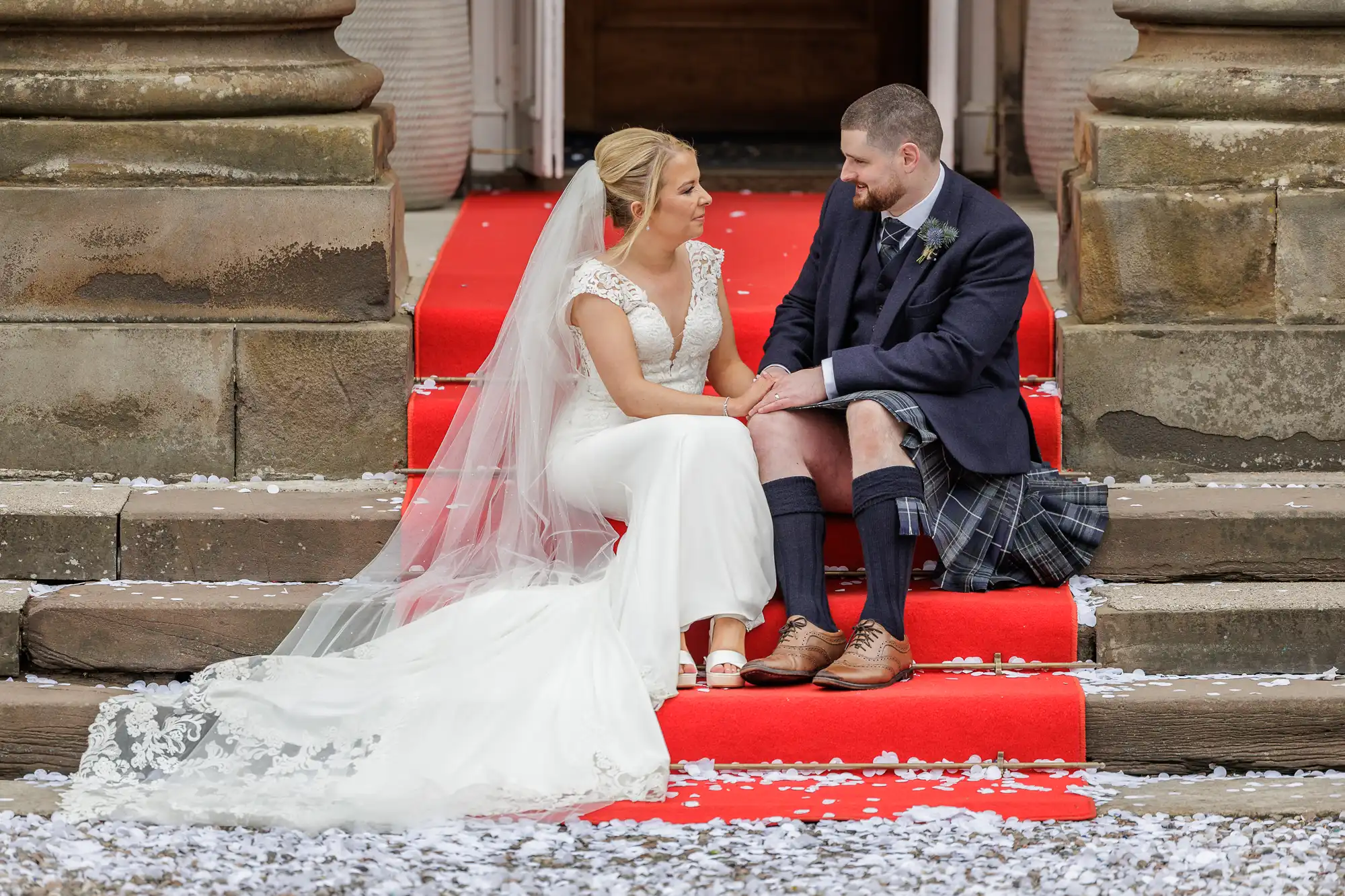 A bride in a white dress and veil sits beside a groom in a kilt and jacket on red-carpeted stone steps. They face each other and hold hands, surrounded by white flower petals, with large stone columns in the background.