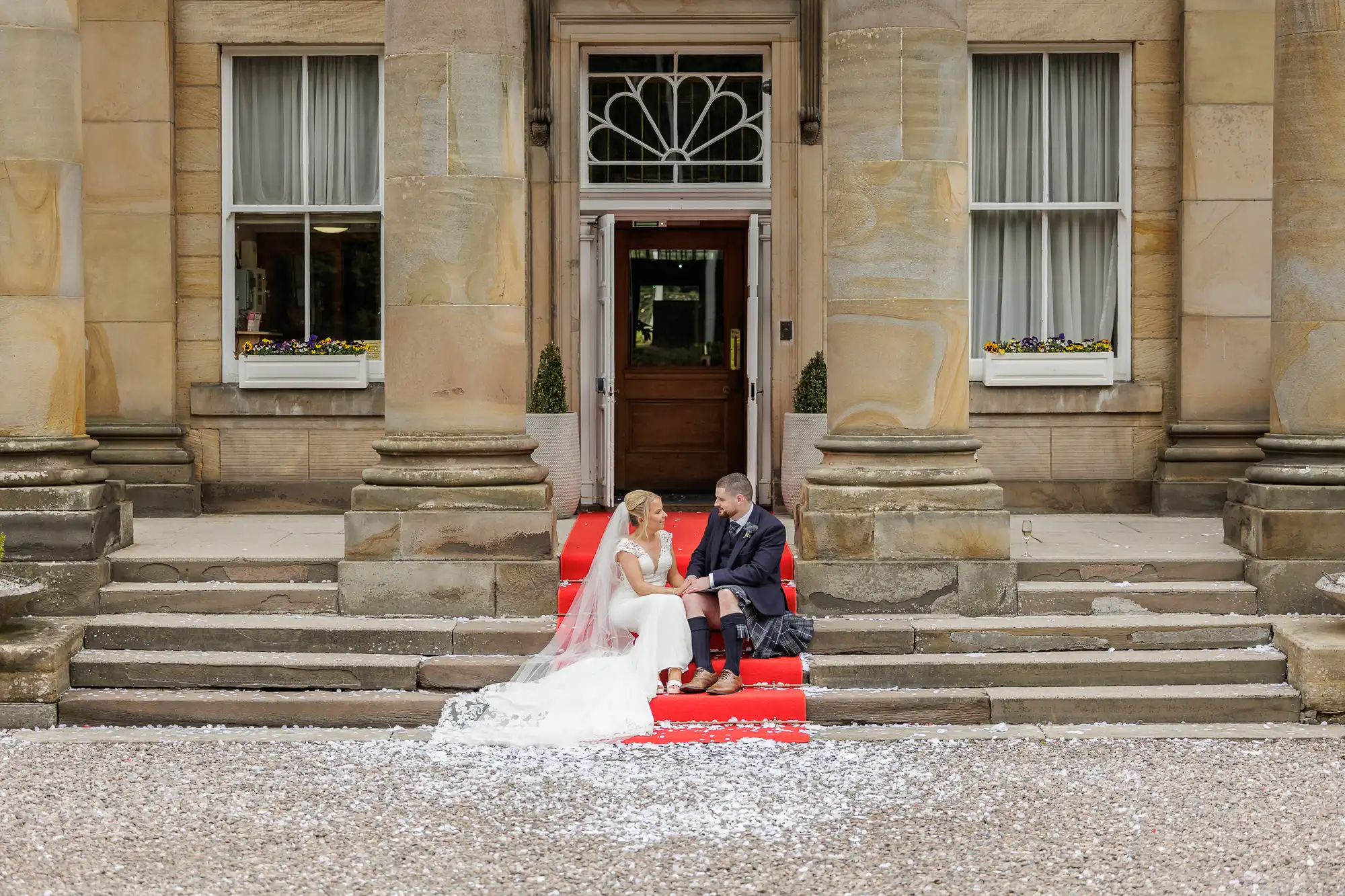 A bride in a white dress and a groom in a dark suit sit together on red-carpeted steps outside a grand stone building with tall columns and large windows, surrounded by scattered white flower petals.