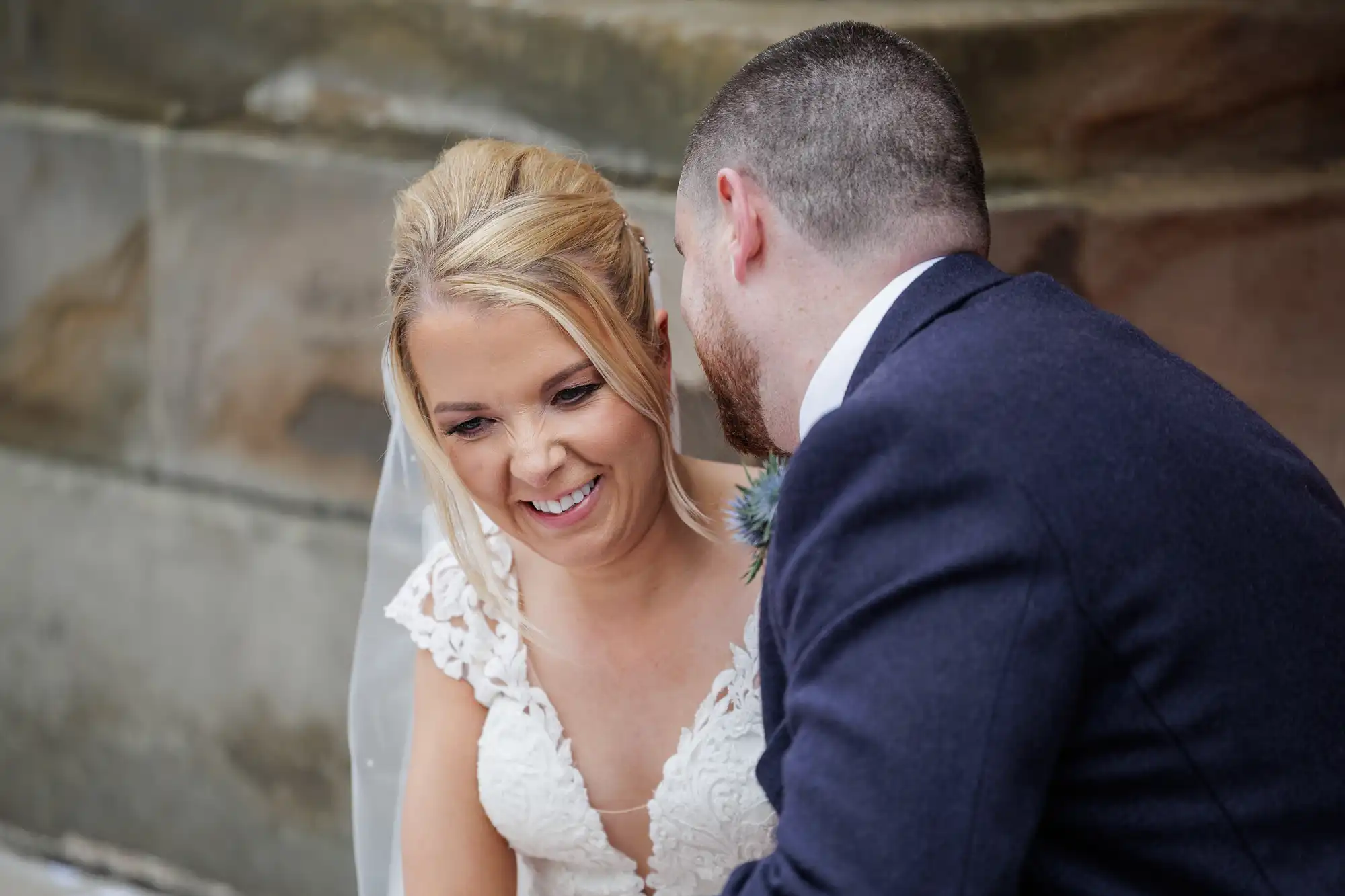 A bride in a white lace dress and veil smiles warmly as a groom in a navy suit leans in close, speaking to her. They are outdoors, standing beside a stone wall, sharing a joyful and intimate moment on their wedding day.