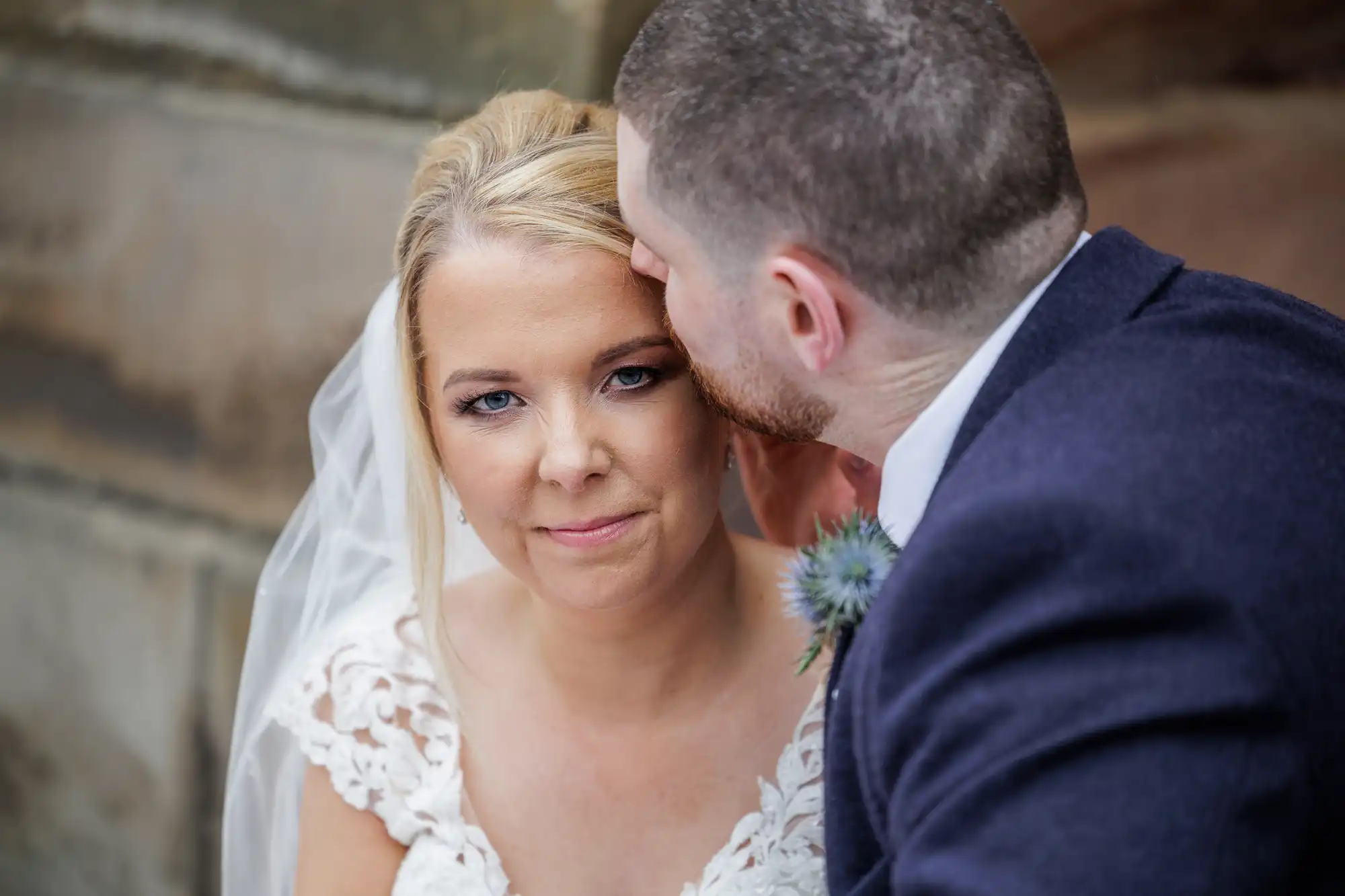 A bride in a white lace dress and veil looks at the camera with a soft smile as a groom in a navy suit gently kisses her forehead. They are positioned close together in front of a blurred stone backdrop.