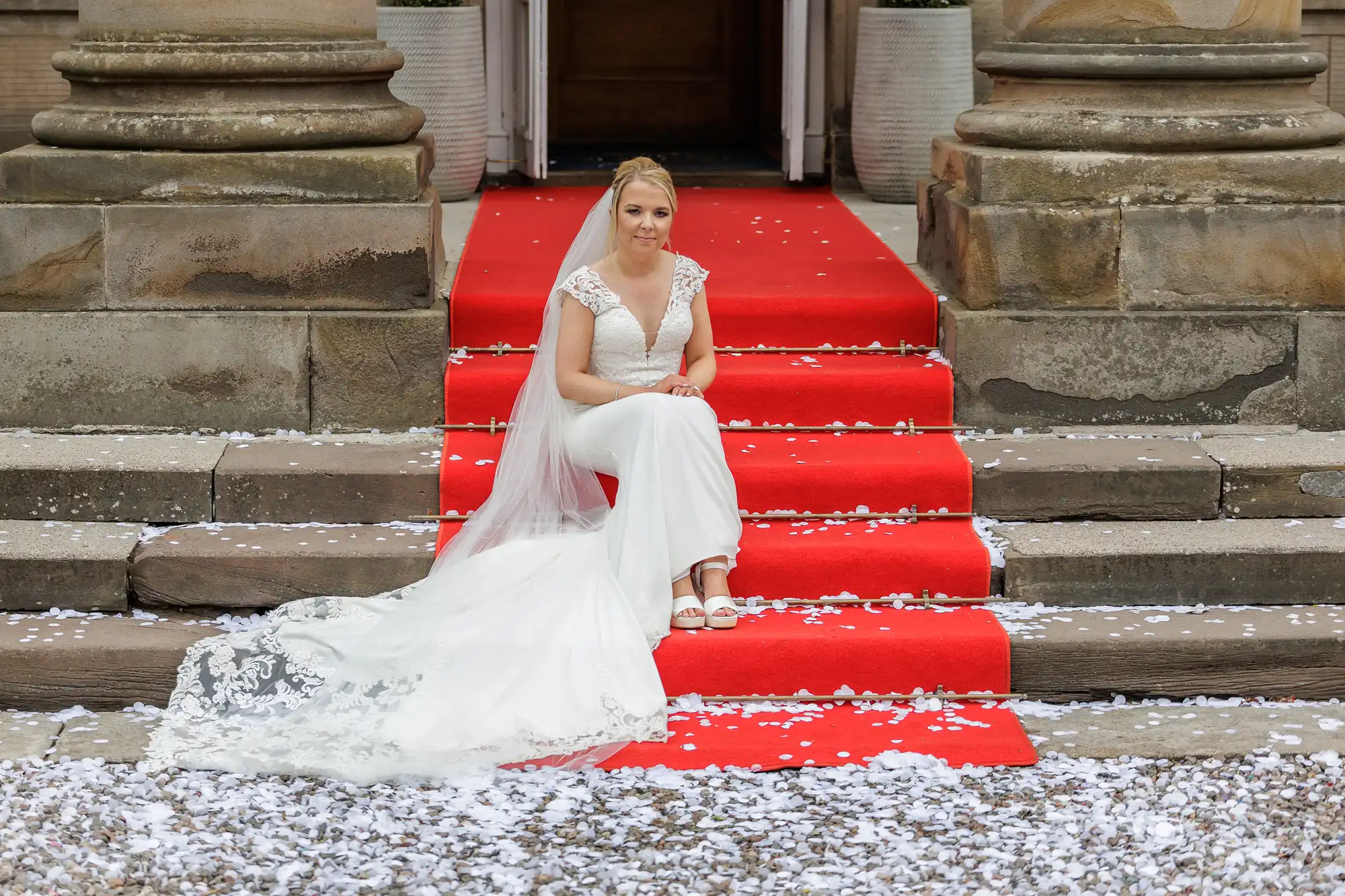 A bride in a white dress and veil sits on stone steps covered with a red carpet. White flower petals are scattered around her. She is facing forward, hands clasped, with large stone columns and potted plants behind her.