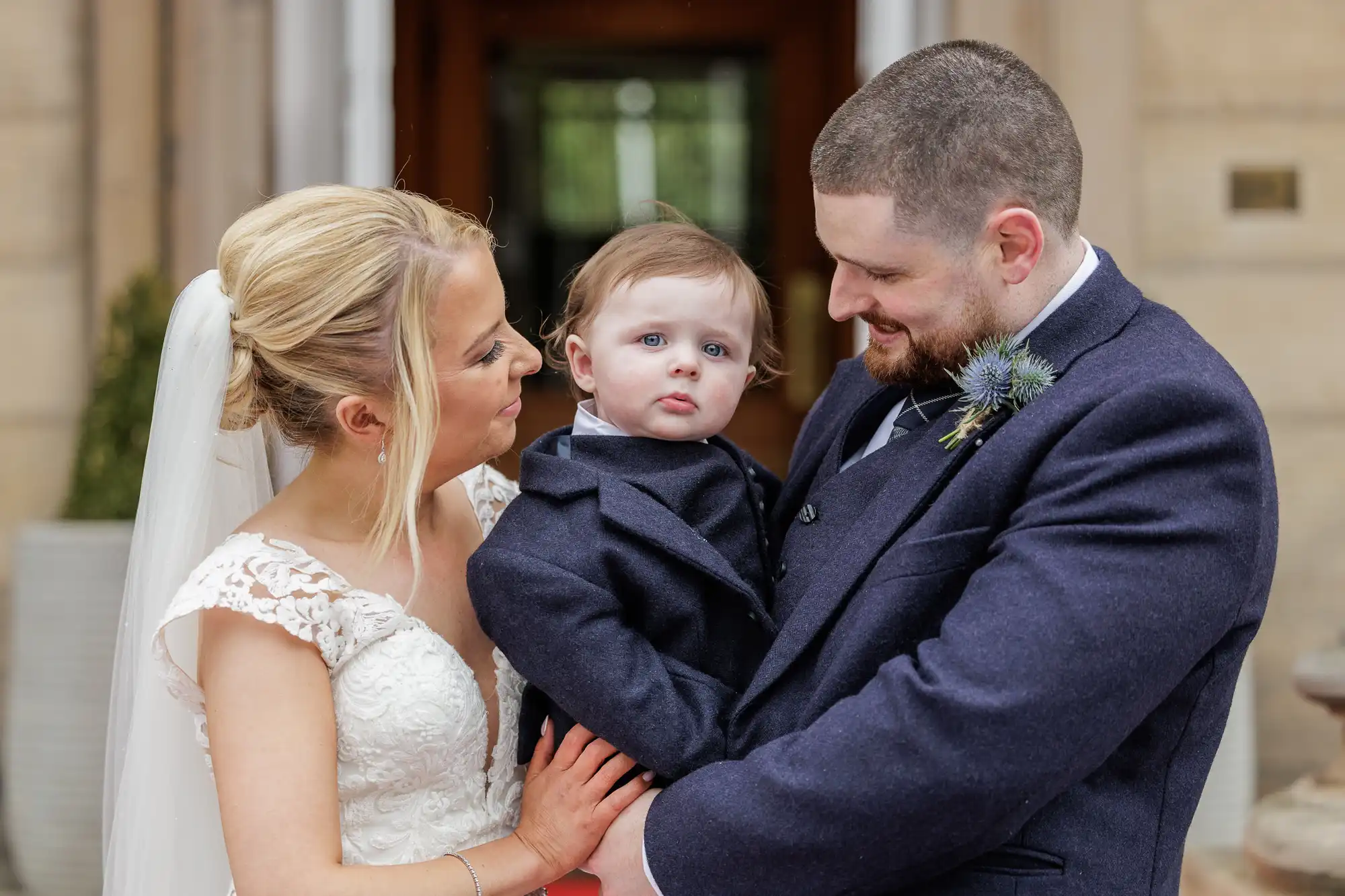 A bride in a white wedding dress and veil stands next to a groom in a navy suit, who is holding a baby in a matching suit. All three are outside a building, smiling and looking at each other. The bride and groom look lovingly at their child.