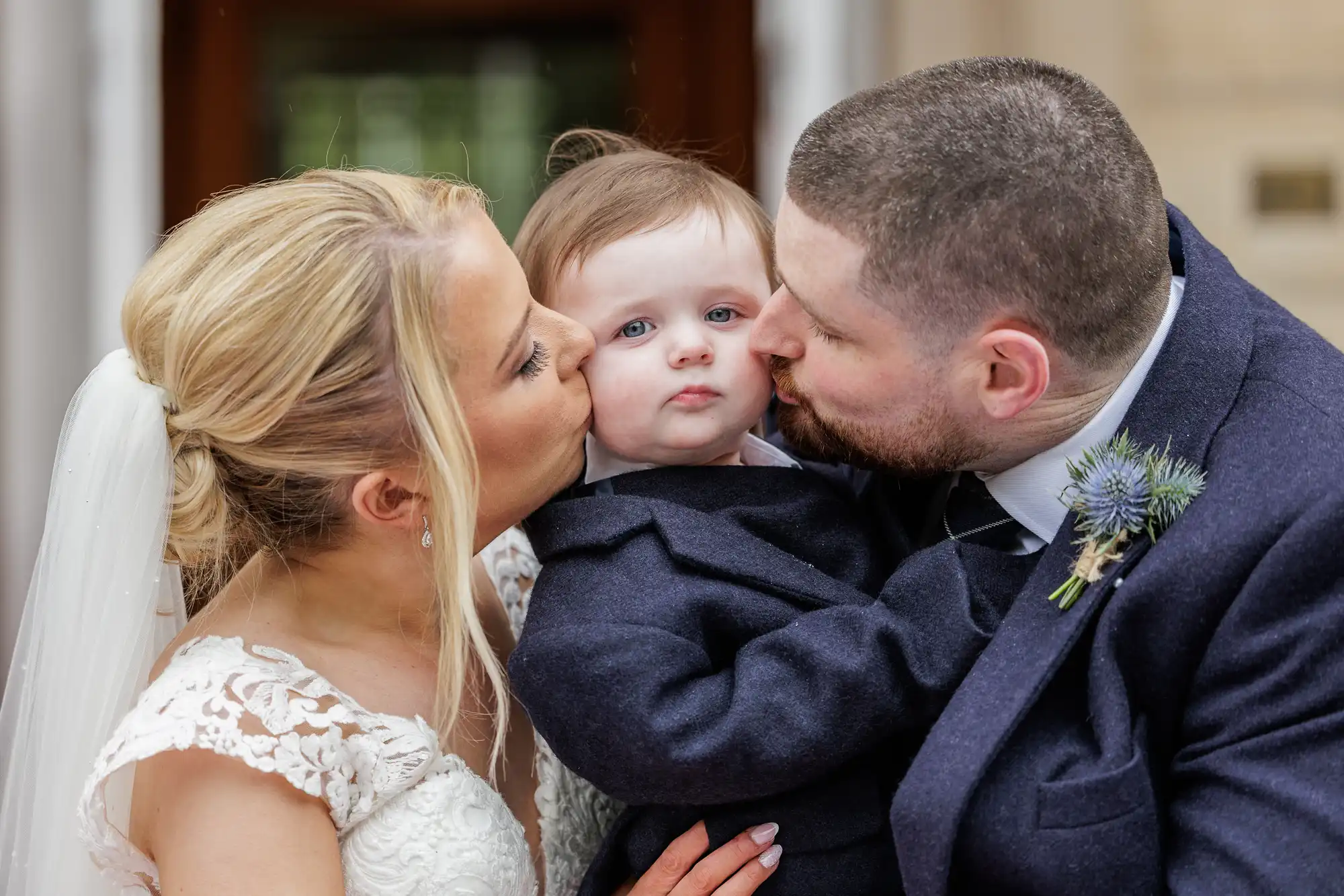 A bride and groom in wedding attire lovingly kiss a young child’s cheeks from either side. The child, dressed in a dark suit, looks directly at the camera with a neutral expression. The background is softly blurred, highlighting the family moment.