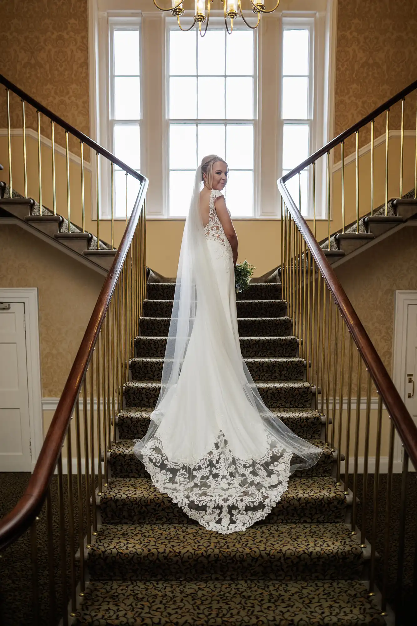 A bride in a white, sleeveless wedding dress with a long lace train and veil stands on a carpeted staircase, looking back over her shoulder. She is framed by gold railings and a large window with natural light behind her.