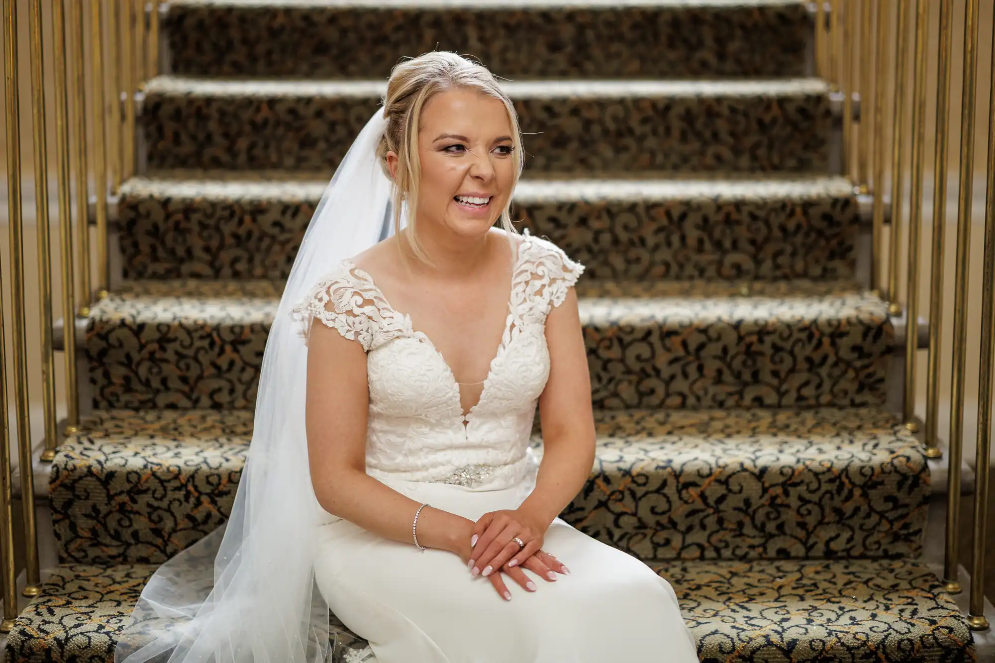 A bride with blonde hair in a low bun and veil sits smiling on patterned carpeted stairs. She wears a white lace wedding dress with cap sleeves and a deep V neckline, and a silver bracelet, holding her hands in her lap. Gold railings frame the stairway.