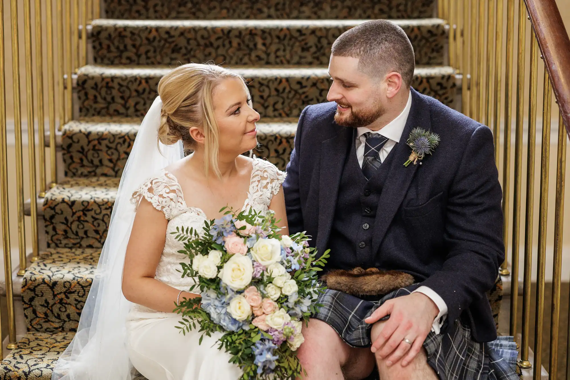 A bride in a white dress holding a pastel bouquet sits beside a groom in a navy jacket, waistcoat, tie, and tartan kilt on carpeted stairs, gazing at each other and smiling. Gold stair rails are visible behind them.