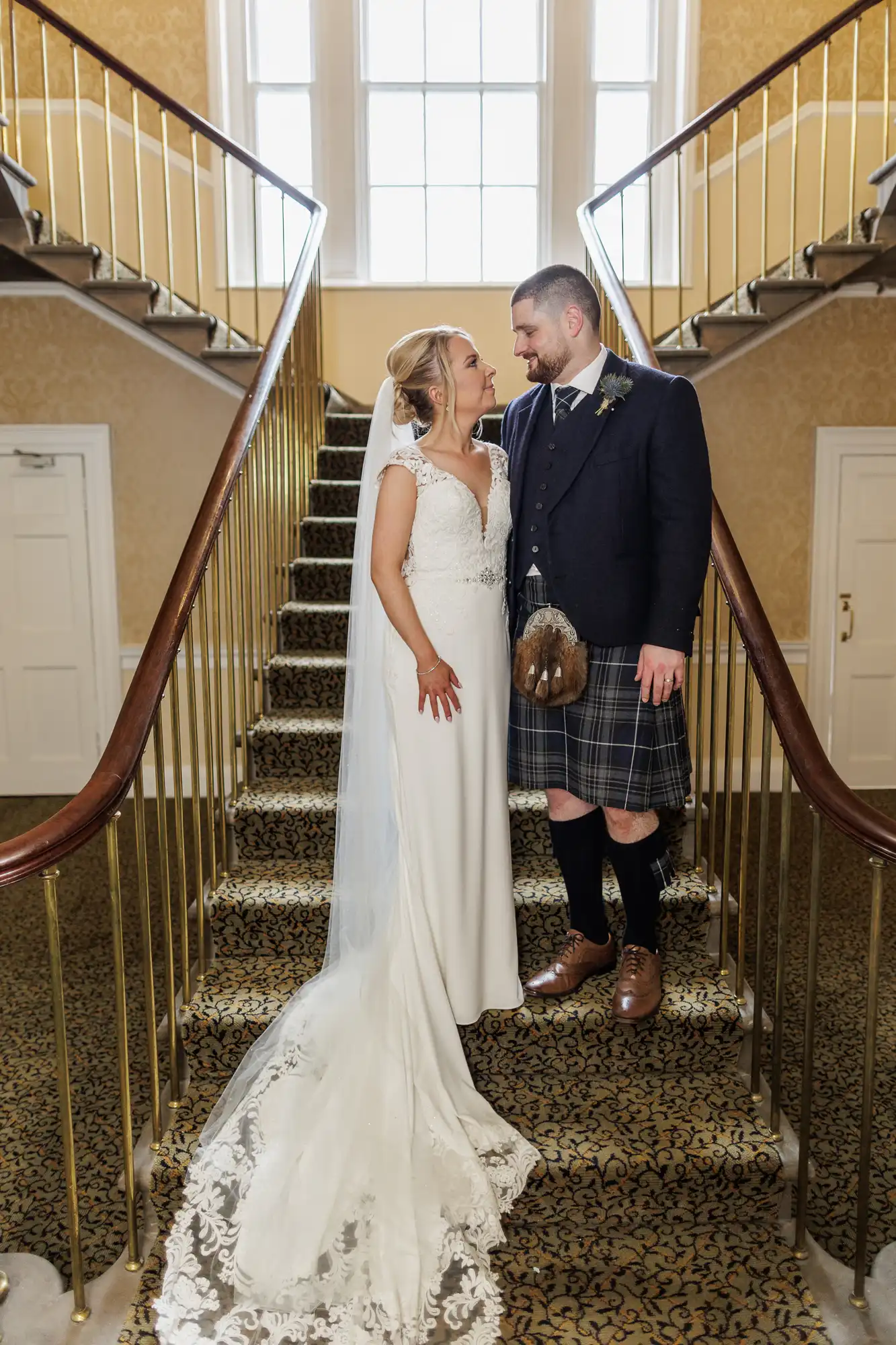 A bride in a white gown with a long train and veil stands on carpeted stairs beside a groom wearing a dark jacket, kilt, and sporran. They face each other, smiling, under tall windows with golden walls and brass railings on both sides.