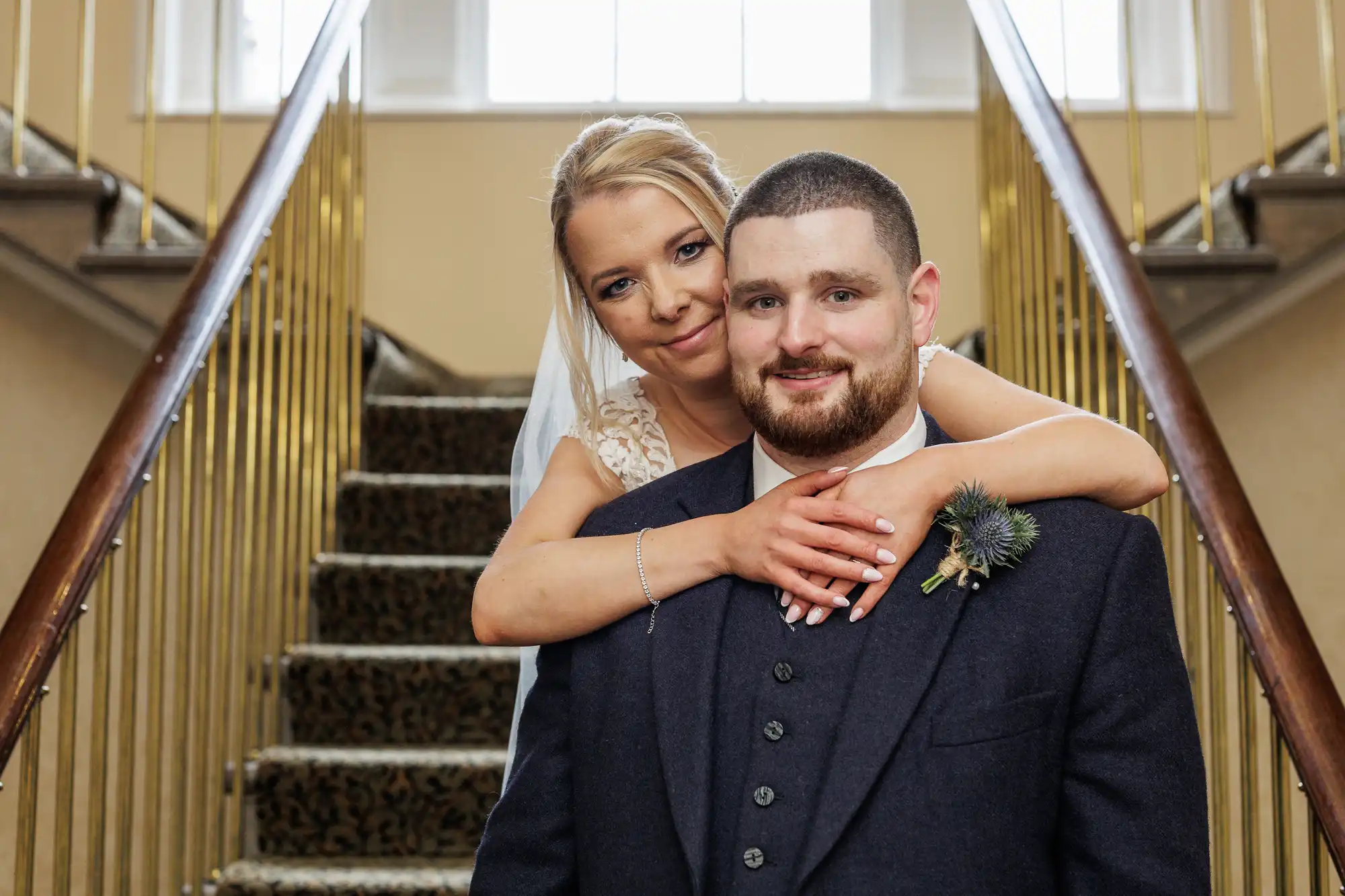 A bride in a white dress embraces a smiling groom in a dark suit from behind as they stand on a carpeted staircase with gold railings. The bride rests her head on his shoulder, and sunlight pours in from windows above the stairs.