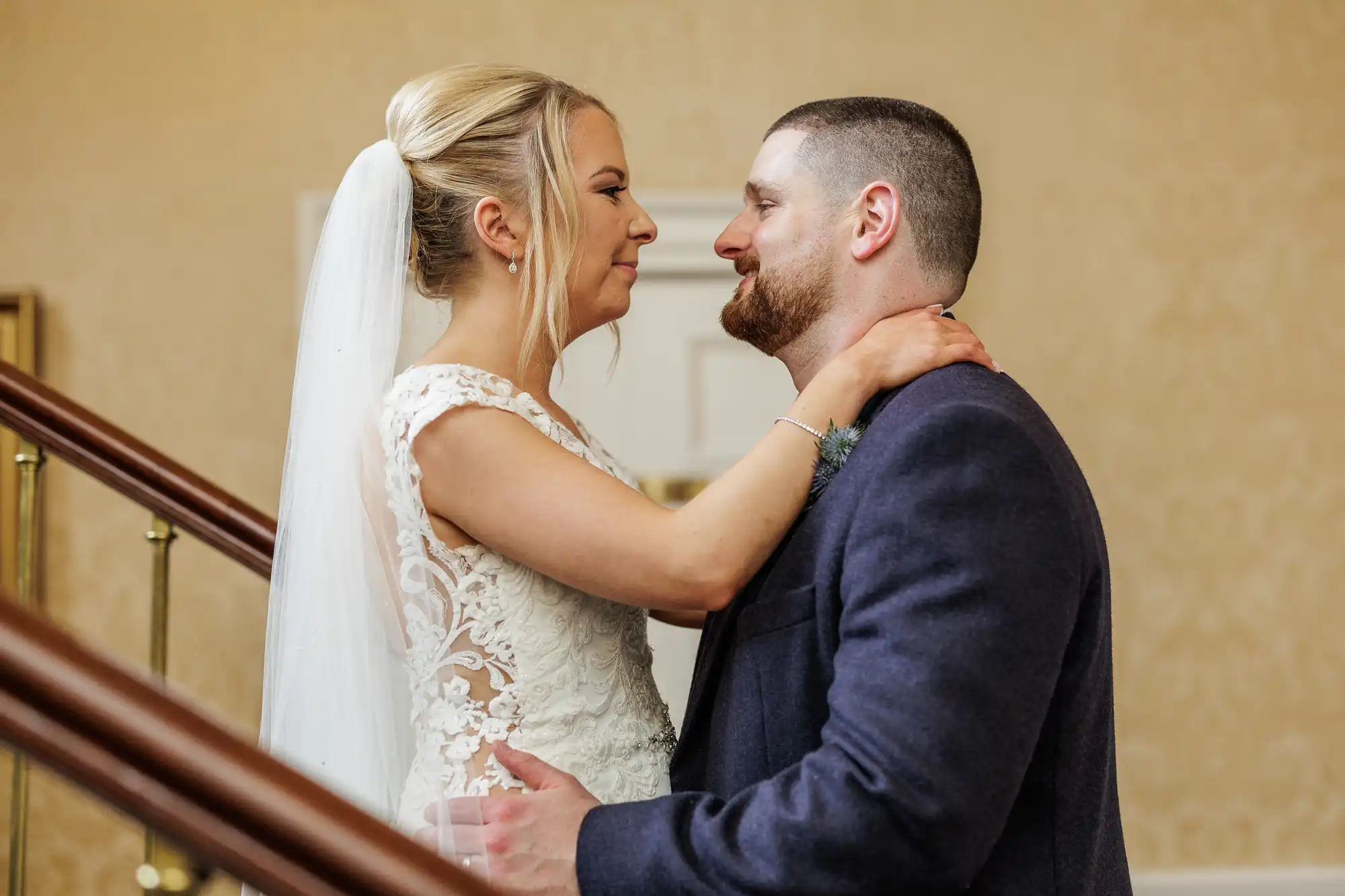 A bride in a white lace dress and veil lovingly embraces a groom in a dark suit. They stand on a staircase, gazing into each other's eyes and smiling, with the bride’s arms around the groom's neck and a blurred neutral background behind them.