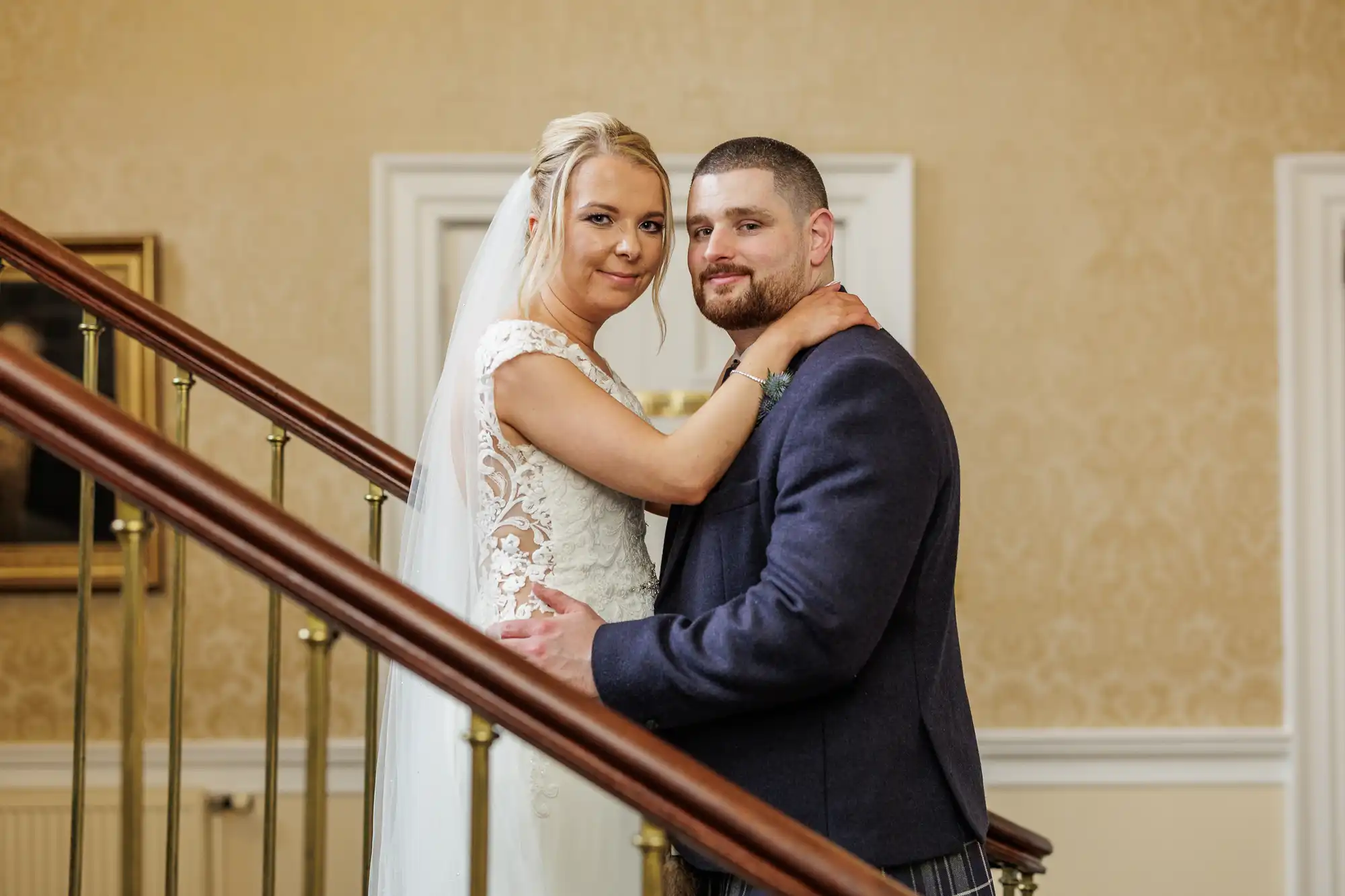 A bride in a white lace gown and veil embraces a groom in a dark suit on an indoor staircase. Both look at the camera, smiling softly. The setting features cream-coloured walls, gold railings, and framed artwork in the background.