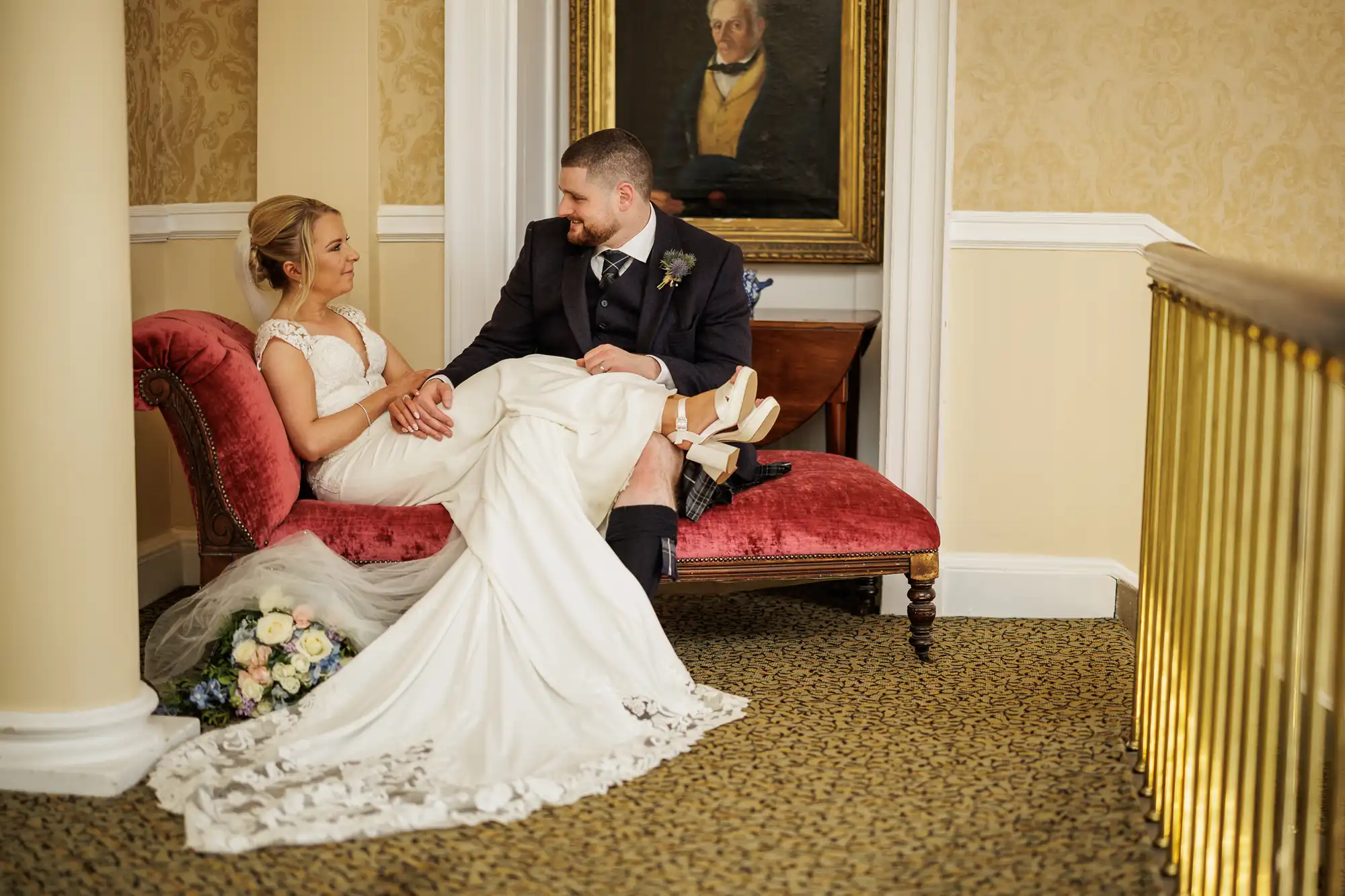 A bride in a white gown reclines on a red chaise longue, holding hands and gazing at a groom in a dark suit. A bouquet rests nearby. They sit in an elegant, gold-accented room with patterned walls and a portrait painting hanging behind them.