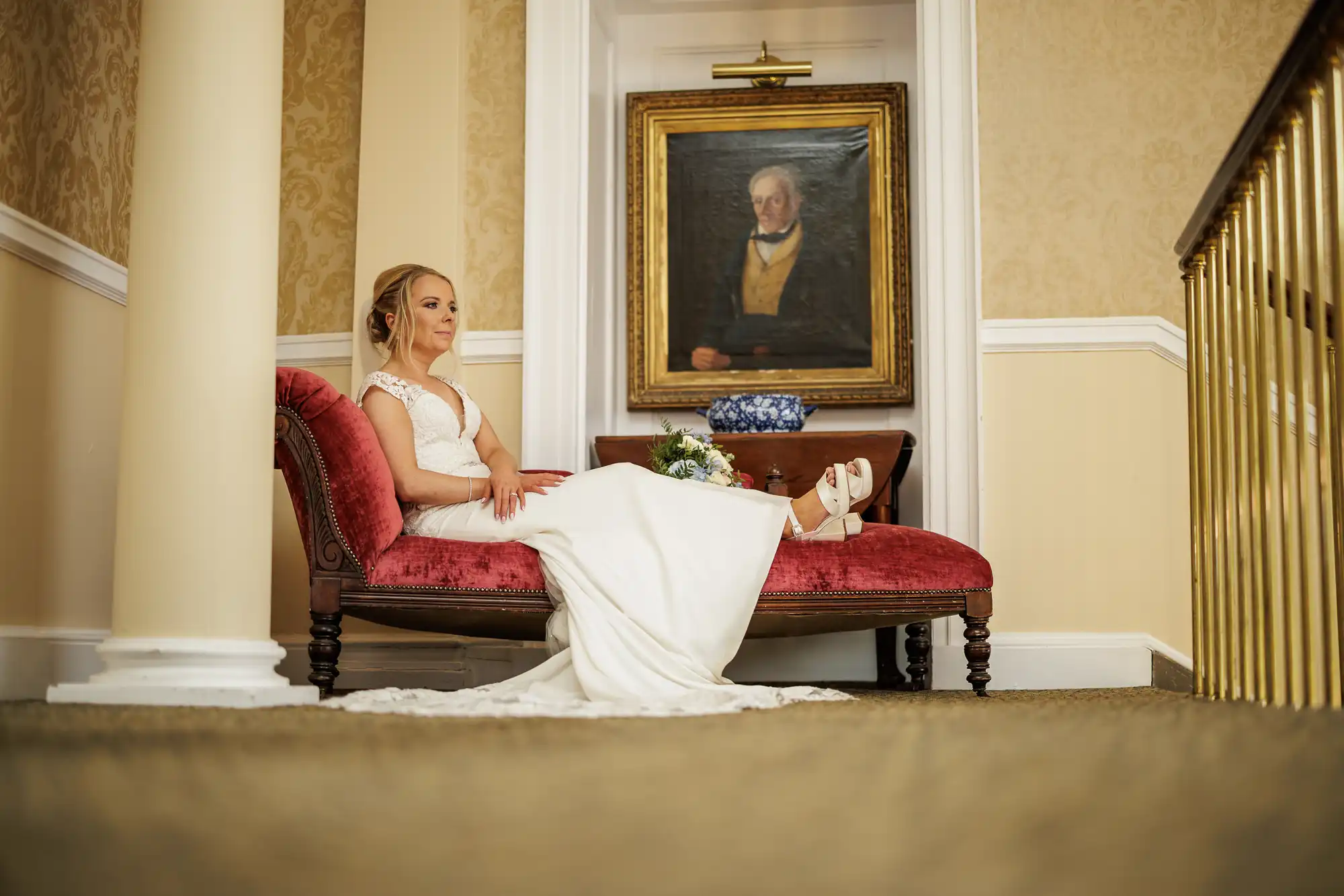 A bride in a white gown sits on a red chaise longue, holding her bouquet. She poses indoors against cream-coloured walls, with a portrait of an older man in historical clothing hanging above her. A blue-and-white vase sits on the table behind her.