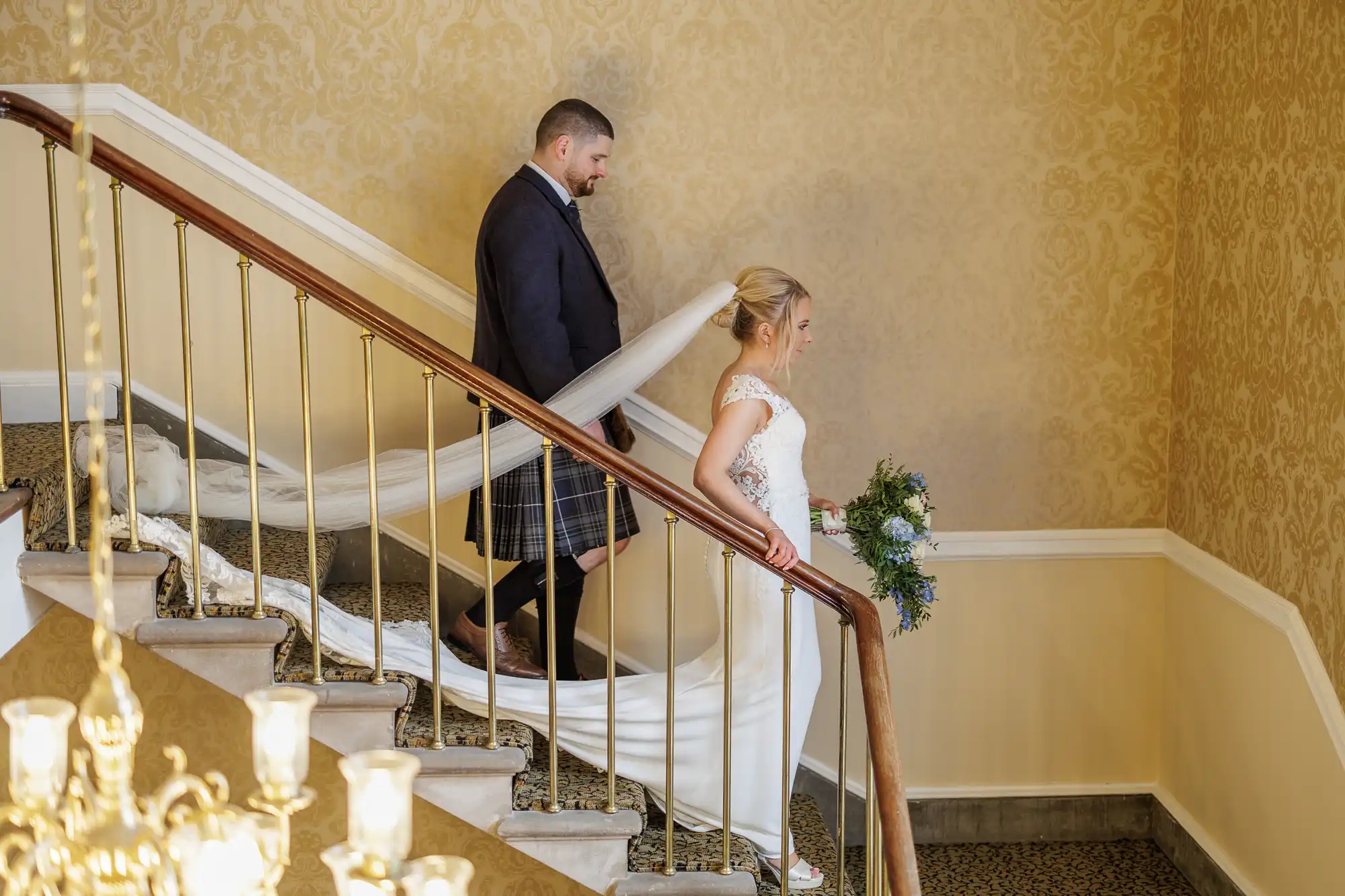 A bride in a white dress holds a bouquet and descends a carpeted staircase with brass railings, followed by a groom in a dark jacket and kilt. Her long veil trails behind her, and the walls are covered in ornate light gold wallpaper.