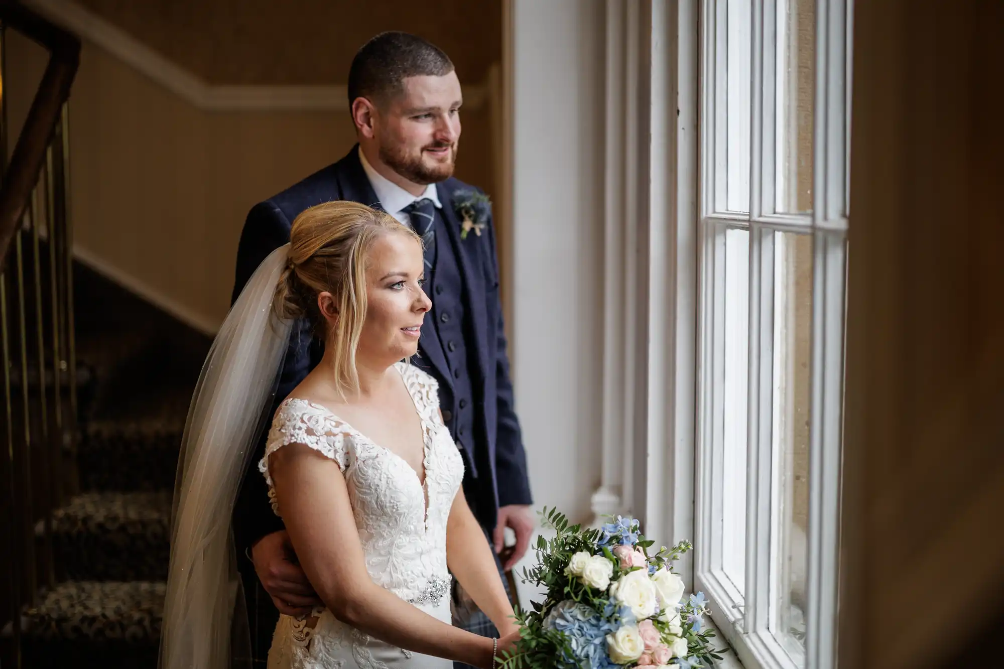 A bride in a white lace gown and veil holds a bouquet of flowers while standing beside a groom in a dark suit. They both look thoughtfully out of a large window, natural light illuminating their faces inside a formal indoor setting.