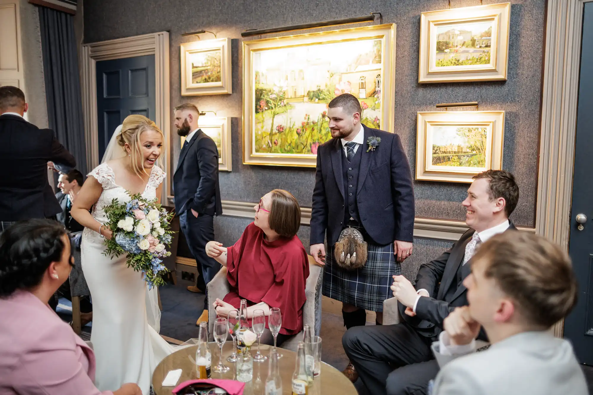 A bride in a white dress and holding a bouquet smiles and leans towards seated guests. A man in a kilt stands beside her. Other guests, dressed formally, chat and smile. The room has blue walls, framed paintings, and drinks on a small round table.