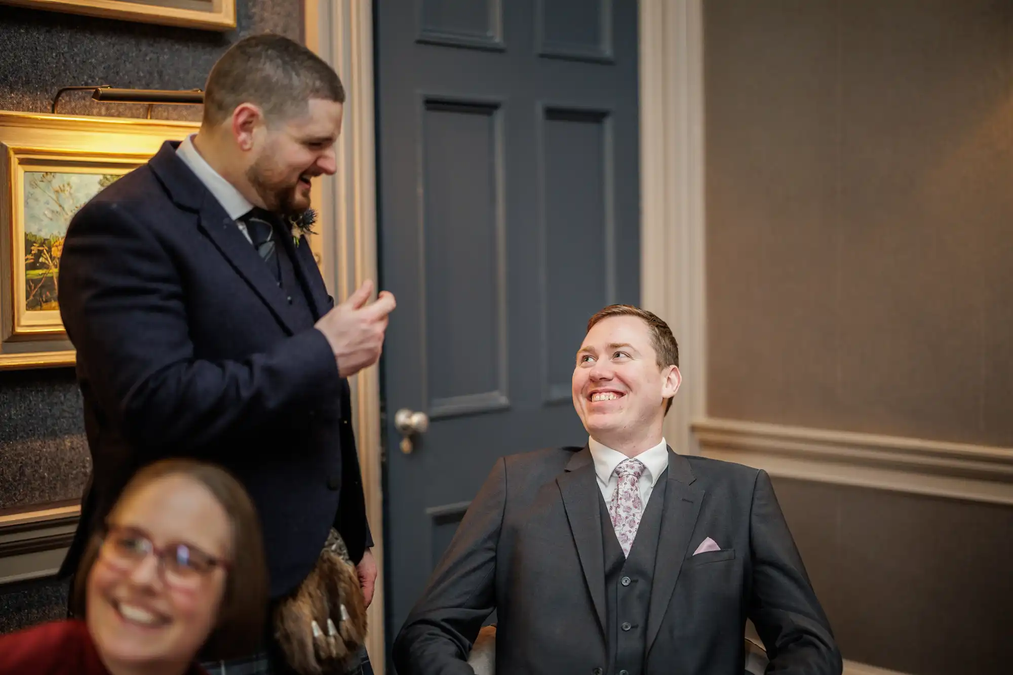 A man in a dark suit stands smiling and gesturing with his hand to another man in a suit who is seated and grinning up at him. A woman with glasses, slightly out of focus, is visible in the foreground, also smiling. Framed art hangs on the wall behind them.