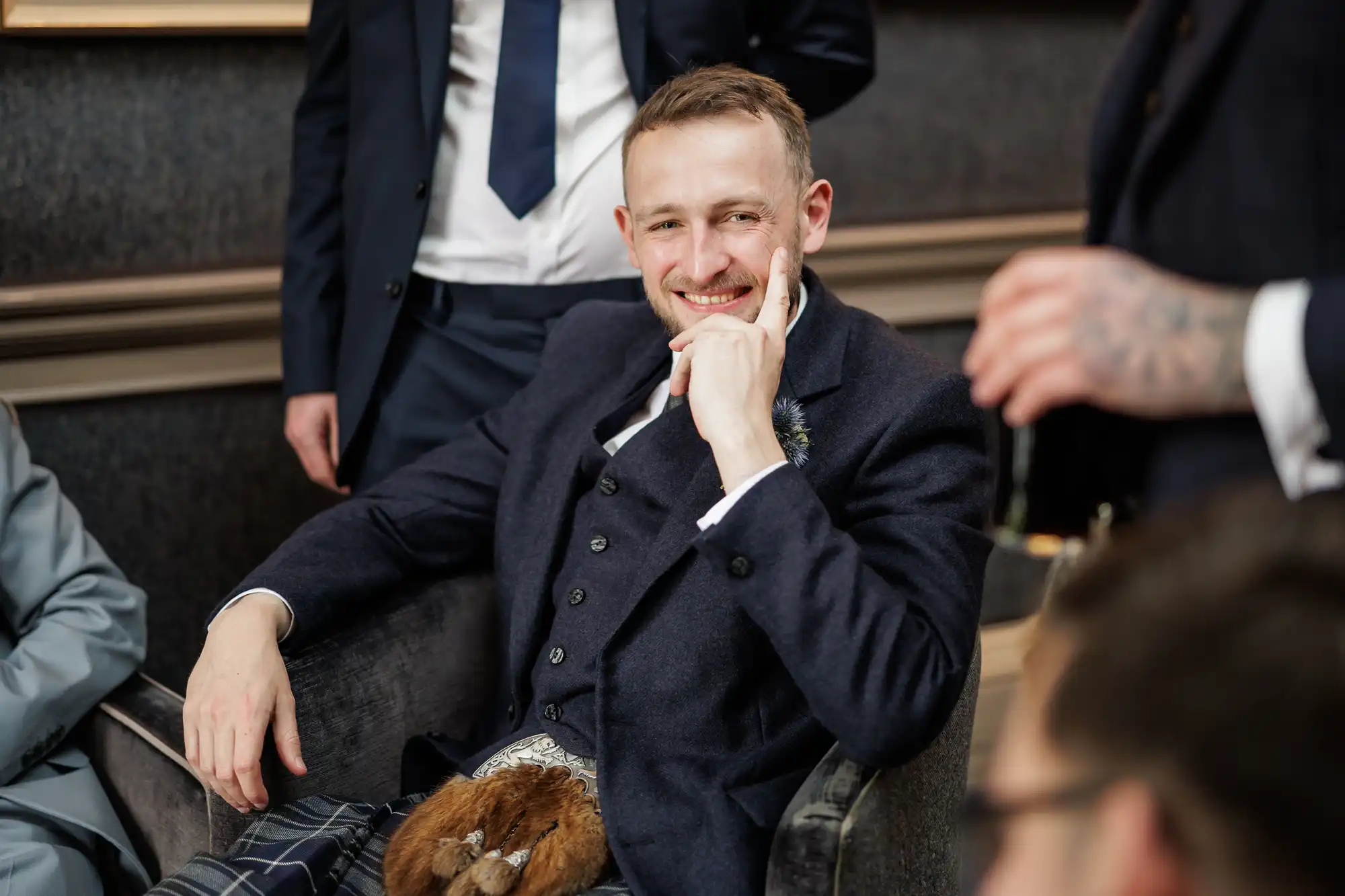 A smiling man in formal Scottish attire, including a dark blazer, waistcoat, and kilt with a sporran, sits in a chair. He rests his head on his hand. Other people in suits stand and sit nearby, partially visible, suggesting a social or formal gathering.