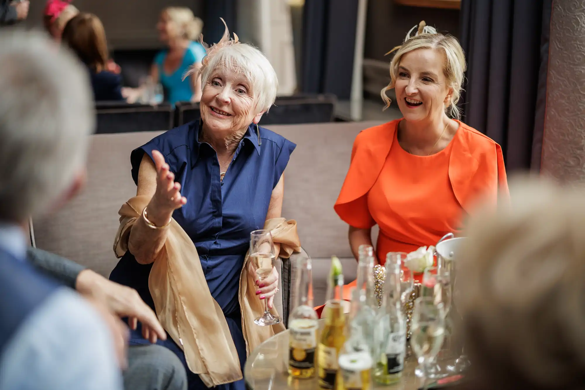 Two older women sit smiling on a sofa at a social gathering, one in a navy dress with a champagne flute, gesturing as she talks, the other in a bright orange dress. Bottles and glasses are on the table, and blurred people are in the background.