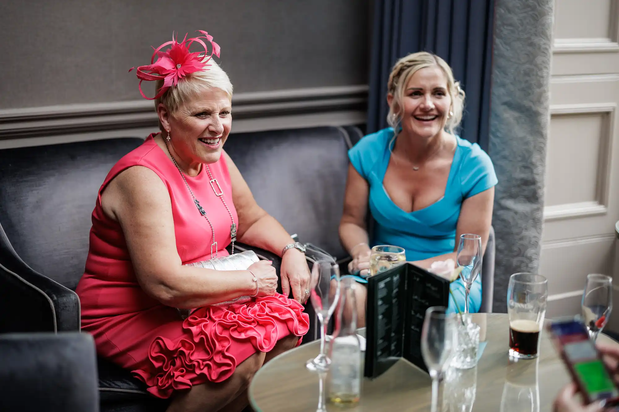 Two smiling women sit on a dark sofa at a social event. One wears a bright pink dress with a matching fascinator, the other a light blue dress. They have drinks in front of them on a glass table, surrounded by empty and half-full glasses.