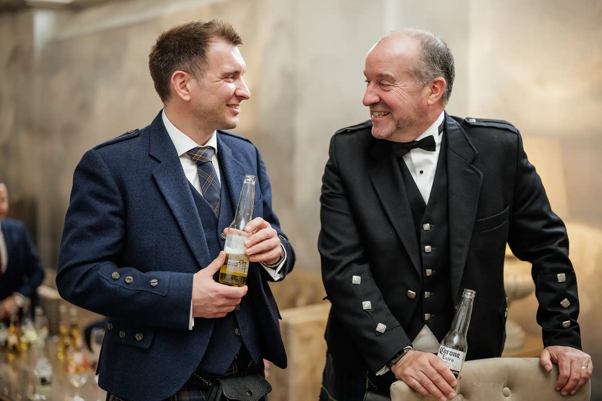 Two men in formal Scottish attire smile at each other while holding beer bottles. One wears a navy suit with a striped tie, the other a black jacket with silver buttons and a bow tie. They stand indoors at an event, surrounded by drink bottles.