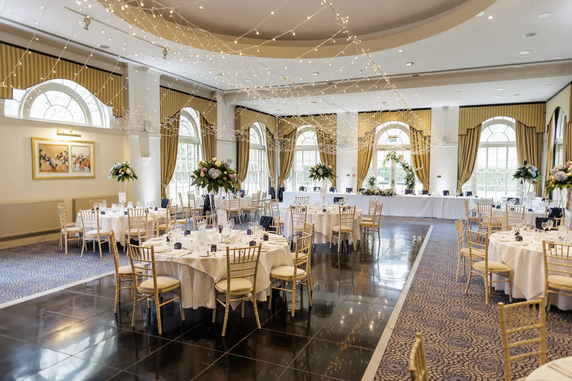 Elegant banquet hall with round tables covered in white cloths, gold chairs, tall floral centrepieces, and tableware. Fairy lights hang from the ceiling. Large arched windows with gold curtains line the walls, letting in natural light.