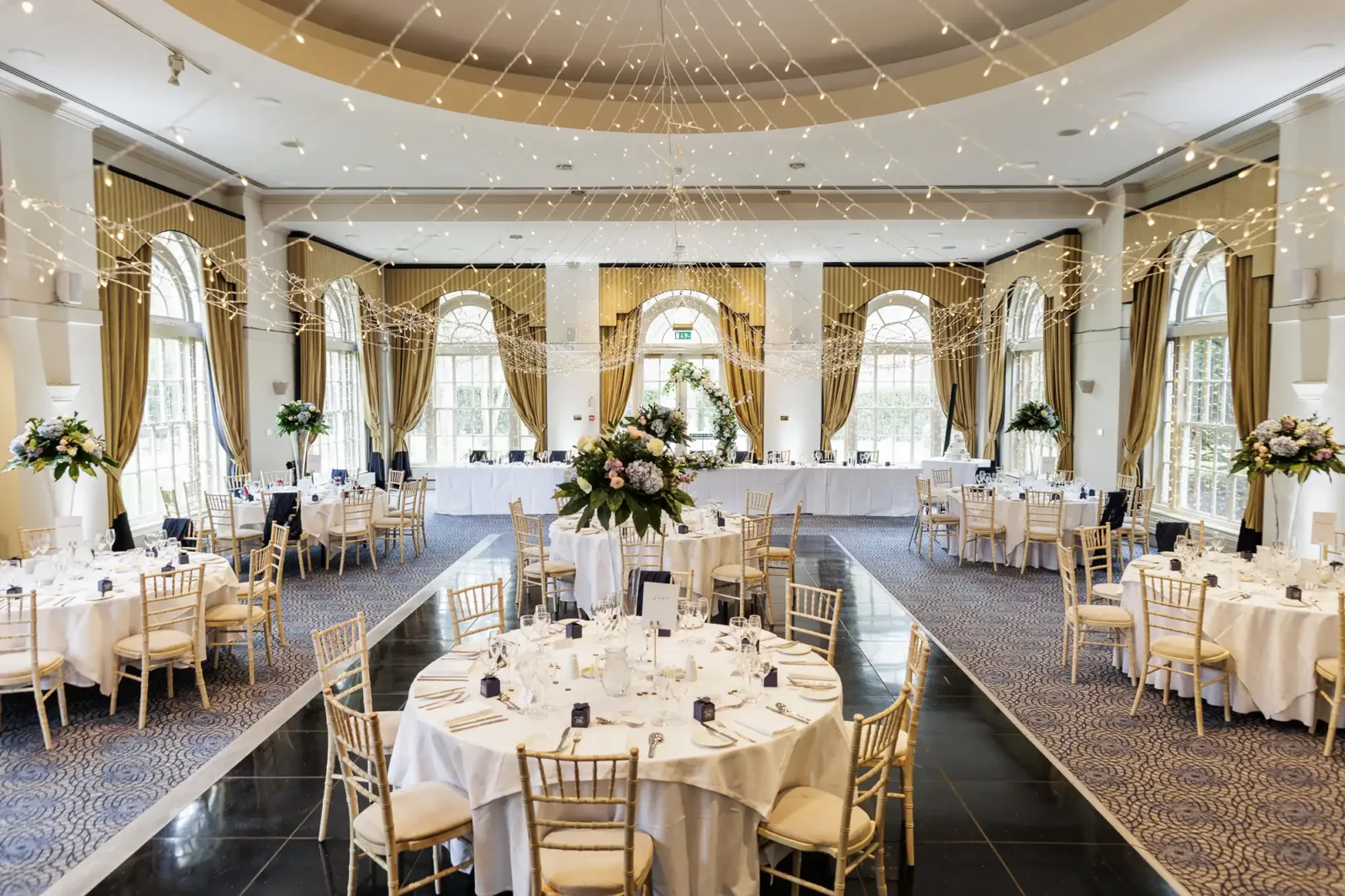 Elegant banqueting hall set for a wedding, with round tables covered in white cloths, gold chairs, floral centrepieces, and hanging fairy lights. Large windows with draped curtains fill the room with natural light. A long top table is at the front.