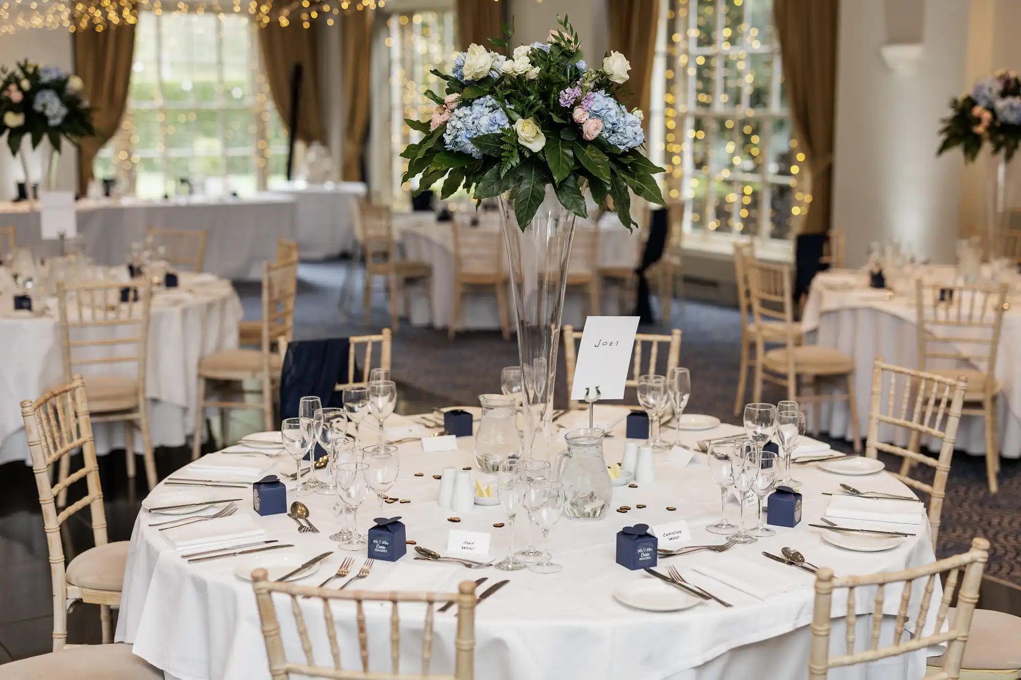 A round banqueting table set for a formal event with white linen, elegant glassware, silver cutlery, blue favour boxes, and a tall floral centrepiece of white, pink, and blue flowers. Other similarly decorated tables and fairy lights are visible in the background.