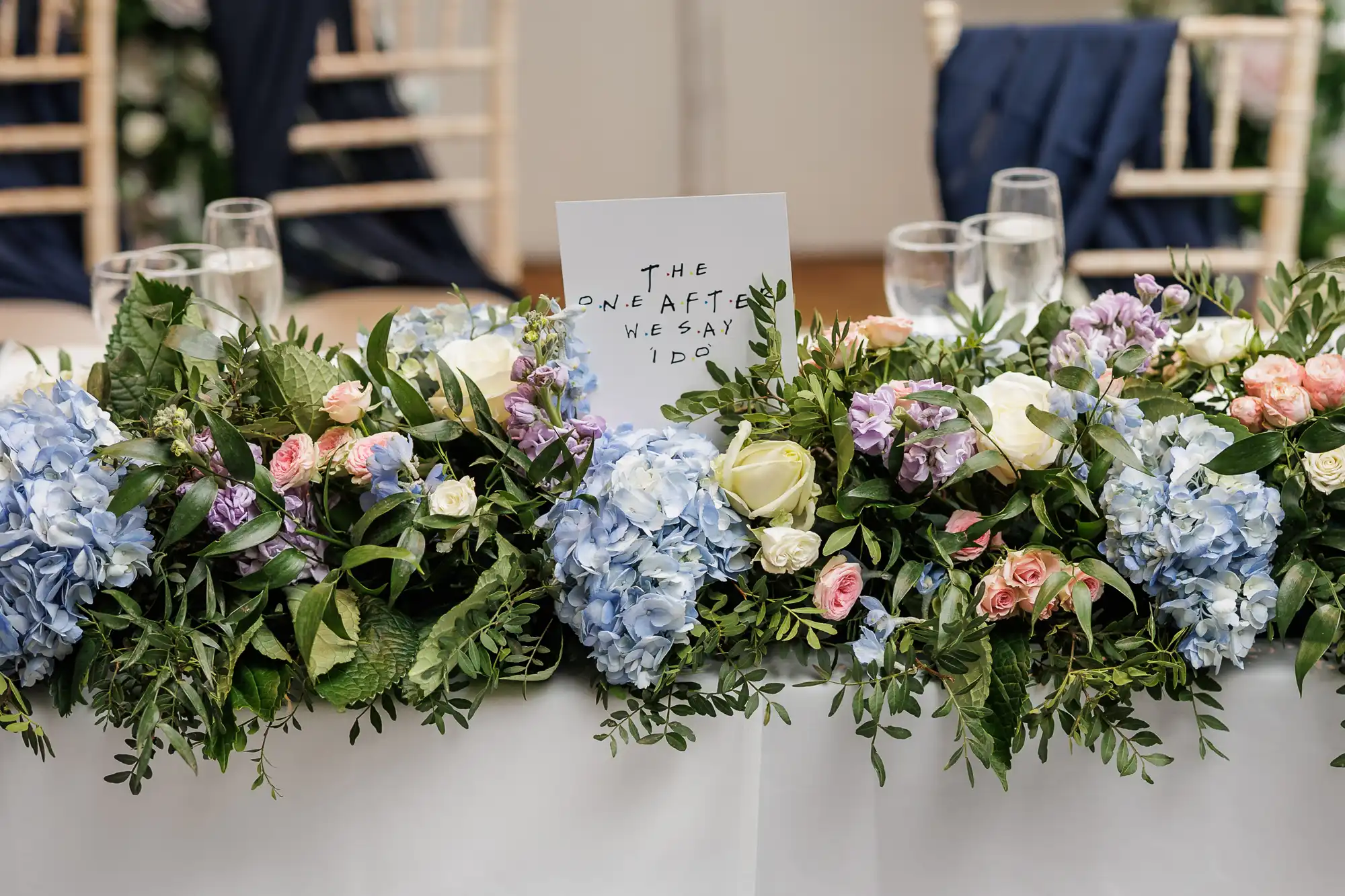A wedding table centrepiece with lush greenery, blue hydrangeas, white roses, and pink flowers. In the centre, a white card reads "THE ONE AFTER WE SAY I DO." In the background are blurred chairs and navy blue fabric accents.