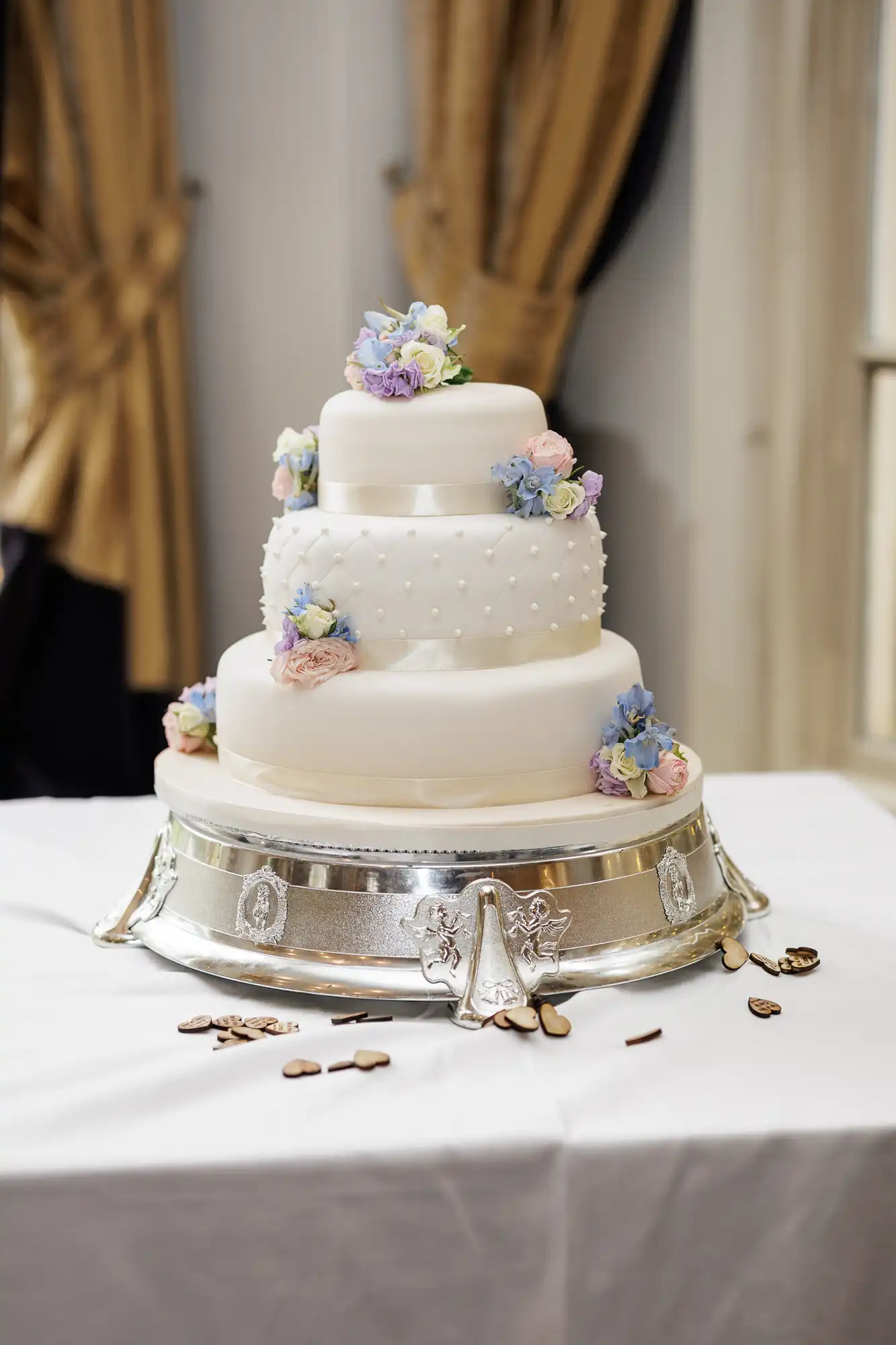 A three-tier white wedding cake decorated with small pearl-like dots and pastel-coloured flowers sits on an ornate silver stand atop a white tablecloth, with a few scattered chocolate pieces on the table. Gold curtains hang in the background.