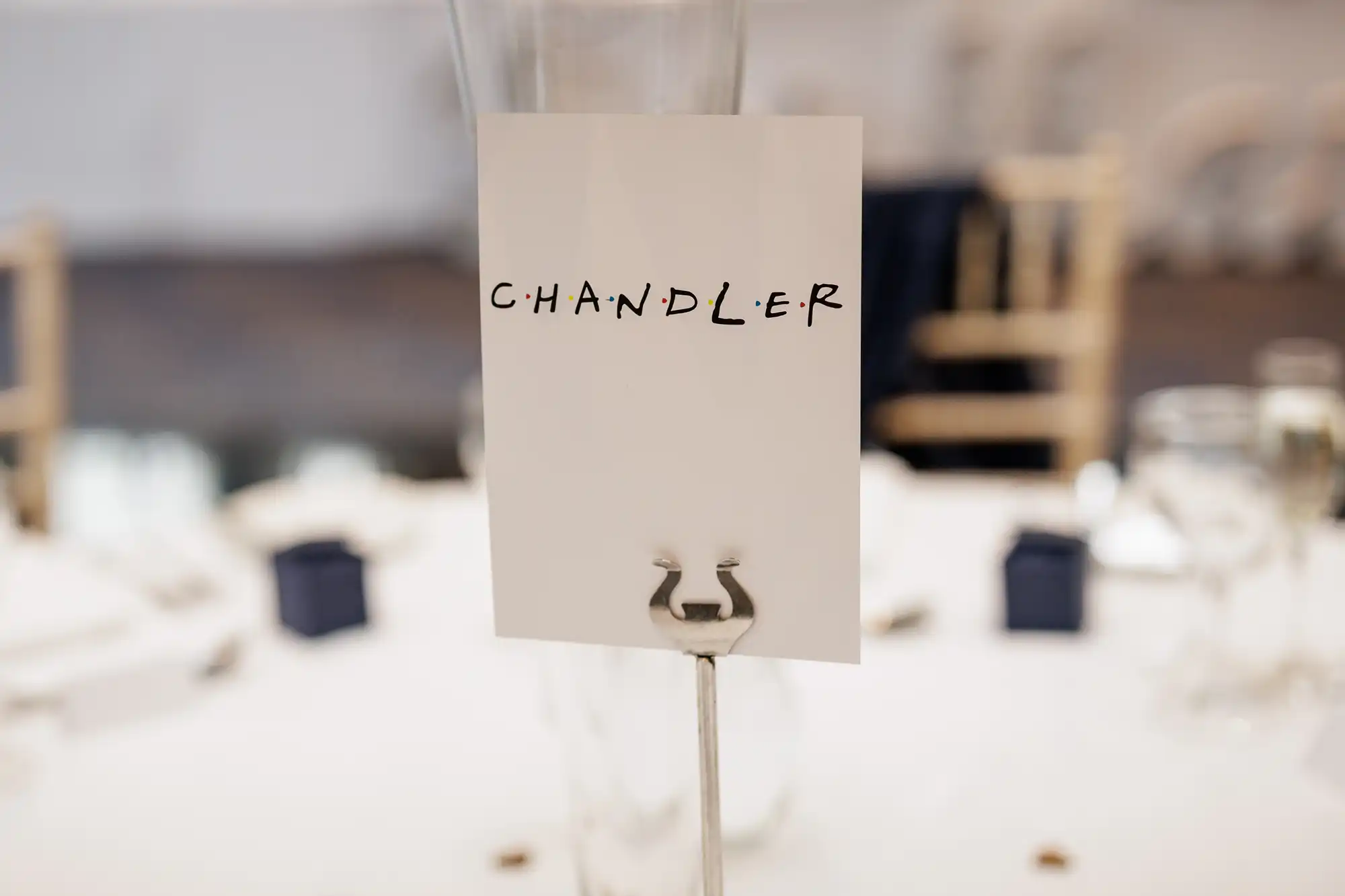 A white card with the word “CHANDLER” in black, playful block letters is held upright by a metal holder on a round, white table. The background shows blurred chairs, glassware, and small navy blue boxes, suggesting an event or celebration.