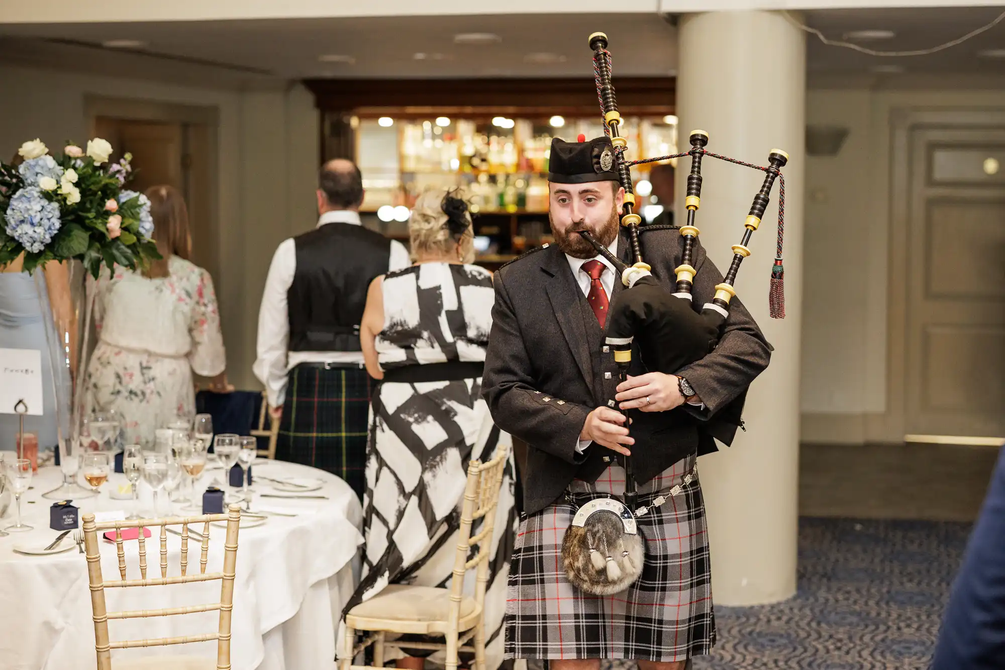 A man in traditional Scottish attire plays bagpipes in a formal dining room. He wears a kilt, jacket, sporran, and hat. Guests in formal clothing are seated at round tables set with glassware and flowers. The setting appears festive and elegant.