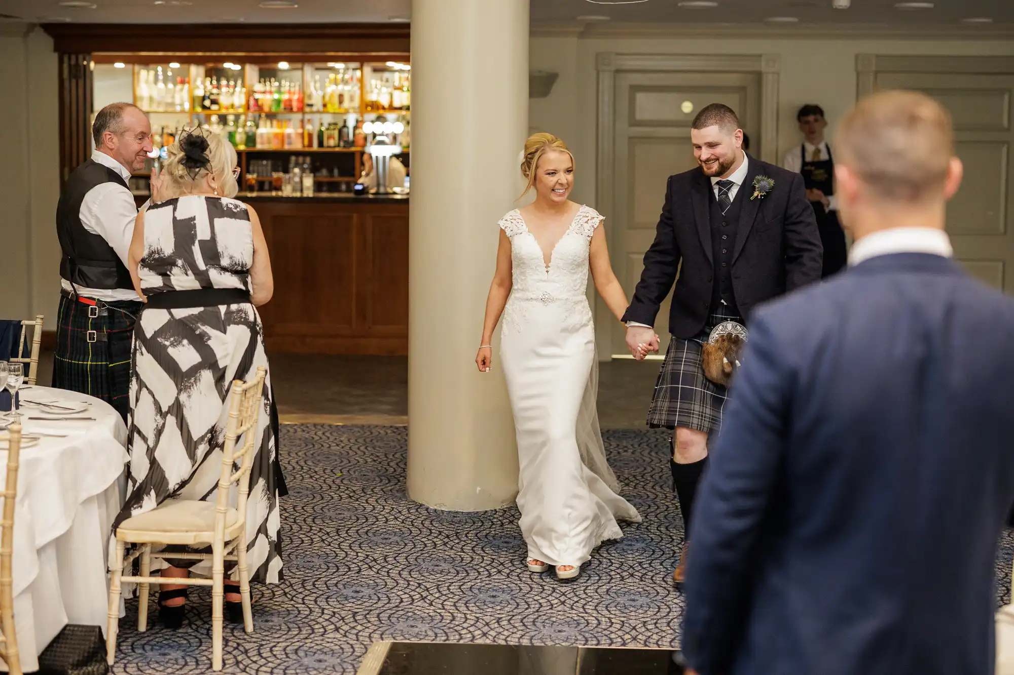 A smiling bride in a white dress and a groom in a kilt walk hand-in-hand into a reception hall. Guests in formal attire watch them enter. The setting features a carpeted floor, a wooden bar with bottles in the background, and a large pillar.