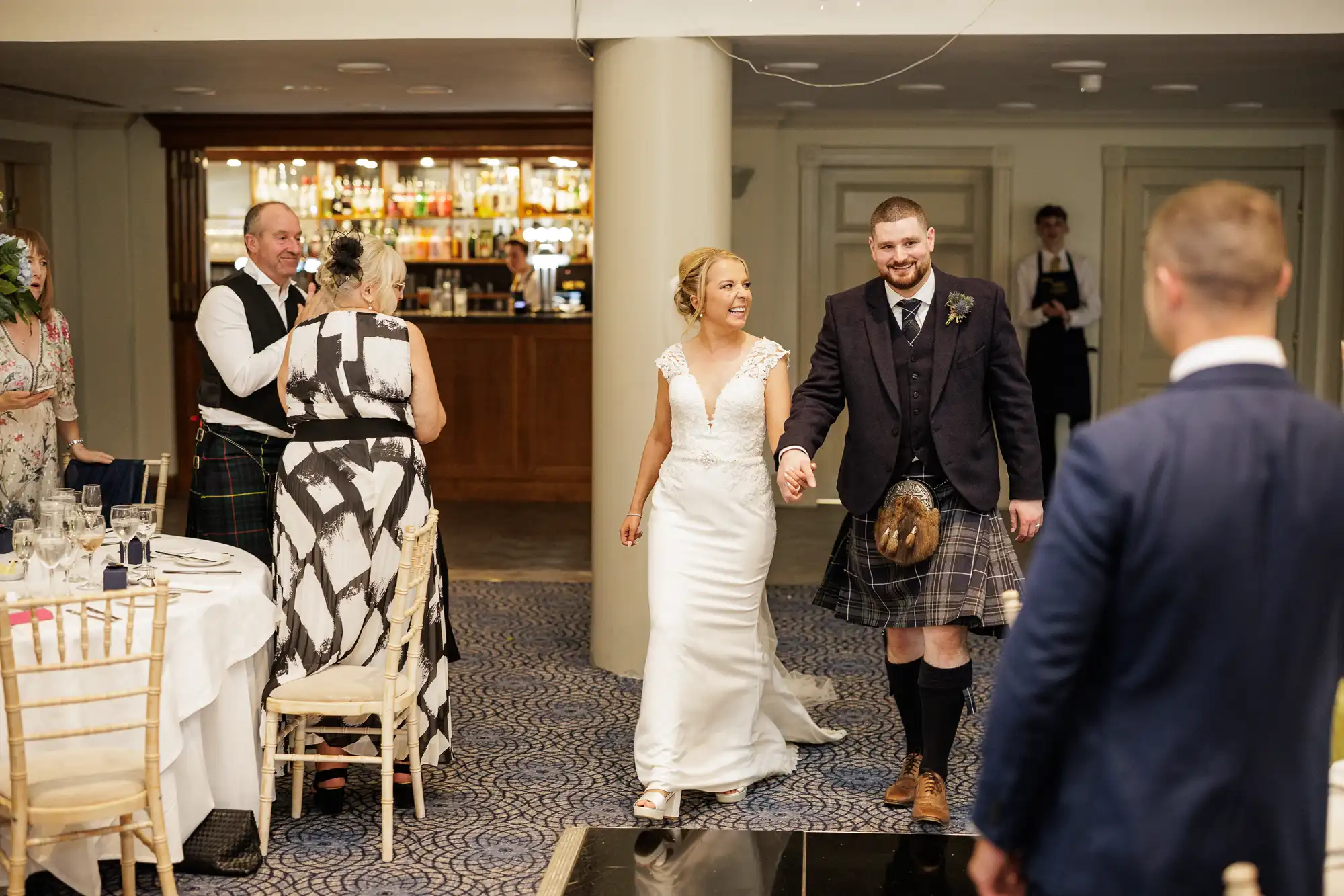 A smiling bride in a white gown and a groom in a kilt and jacket hold hands, entering a reception room. Guests stand and clap near round tables with white tablecloths. A bar with bottles is visible in the background; the scene is cheerful and celebratory.