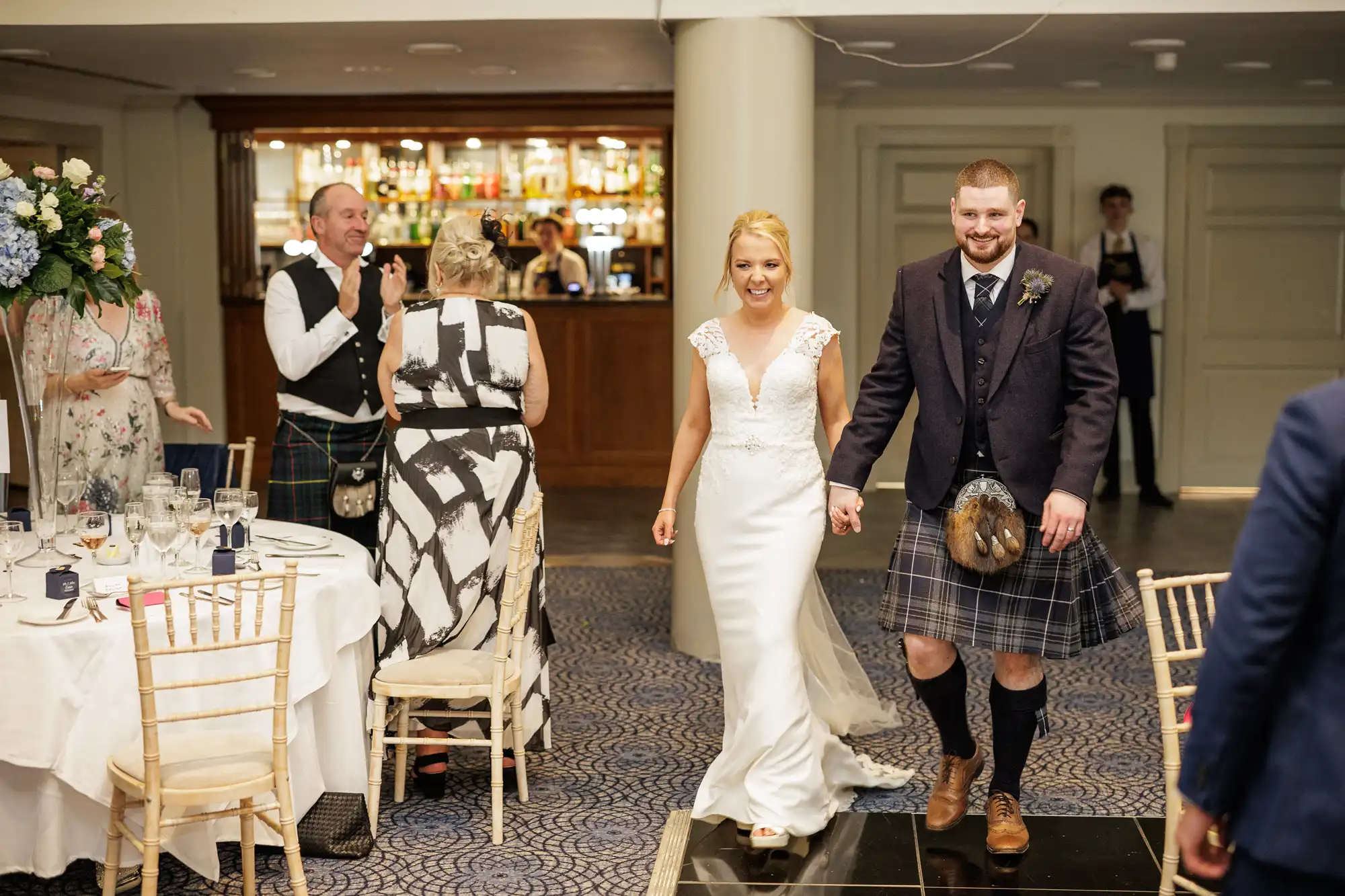 A bride in a white gown and a groom in a kilt and dark jacket smile as they enter a reception area. Guests at round tables with white cloths clap and look on. The background shows a bar with bottles and glasses, and the atmosphere is festive.
