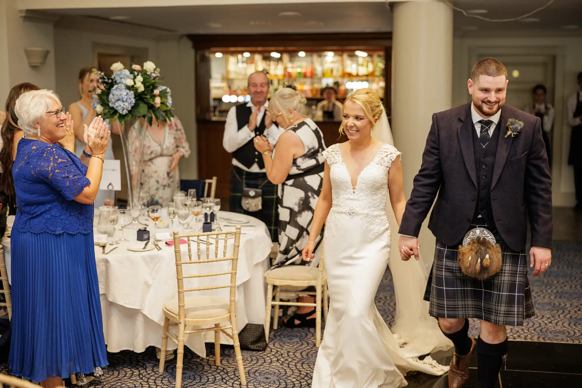 A bride in a white dress and a groom in a kilt and jacket enter a wedding reception, holding hands and smiling. Guests at round tables applaud and smile. A woman in a blue dress stands clapping in the foreground. The setting is elegant with white and blue decor.
