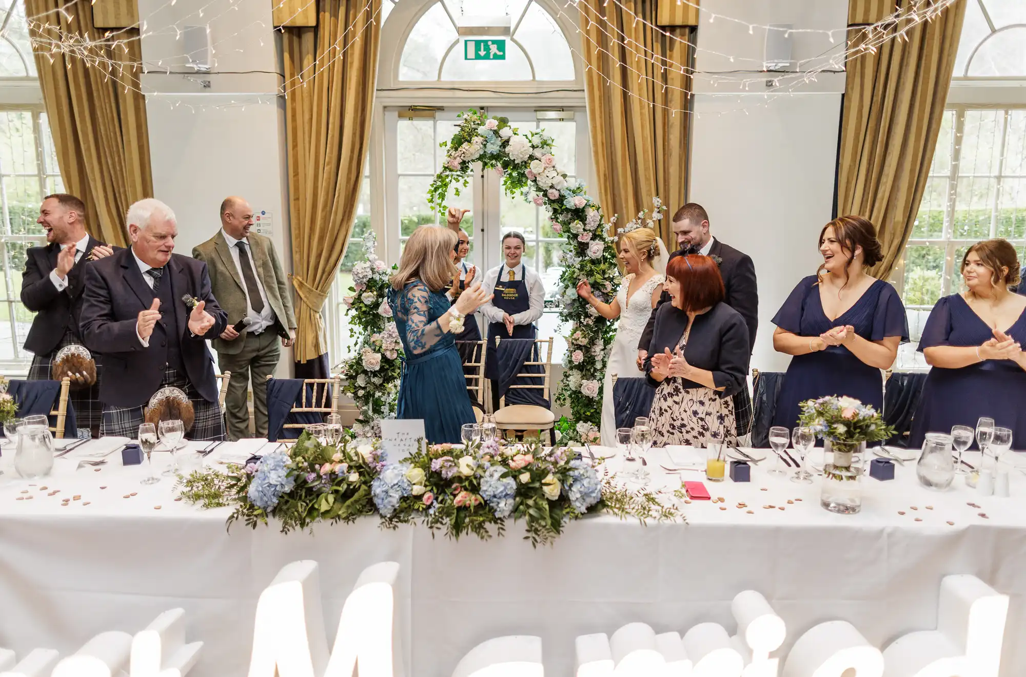 A bride in a blue dress stands at a wedding reception table decorated with flowers and greenery. Guests around her, dressed formally, are smiling, applauding, and looking at her. Fairy lights hang above, and large windows with gold curtains fill the background.
