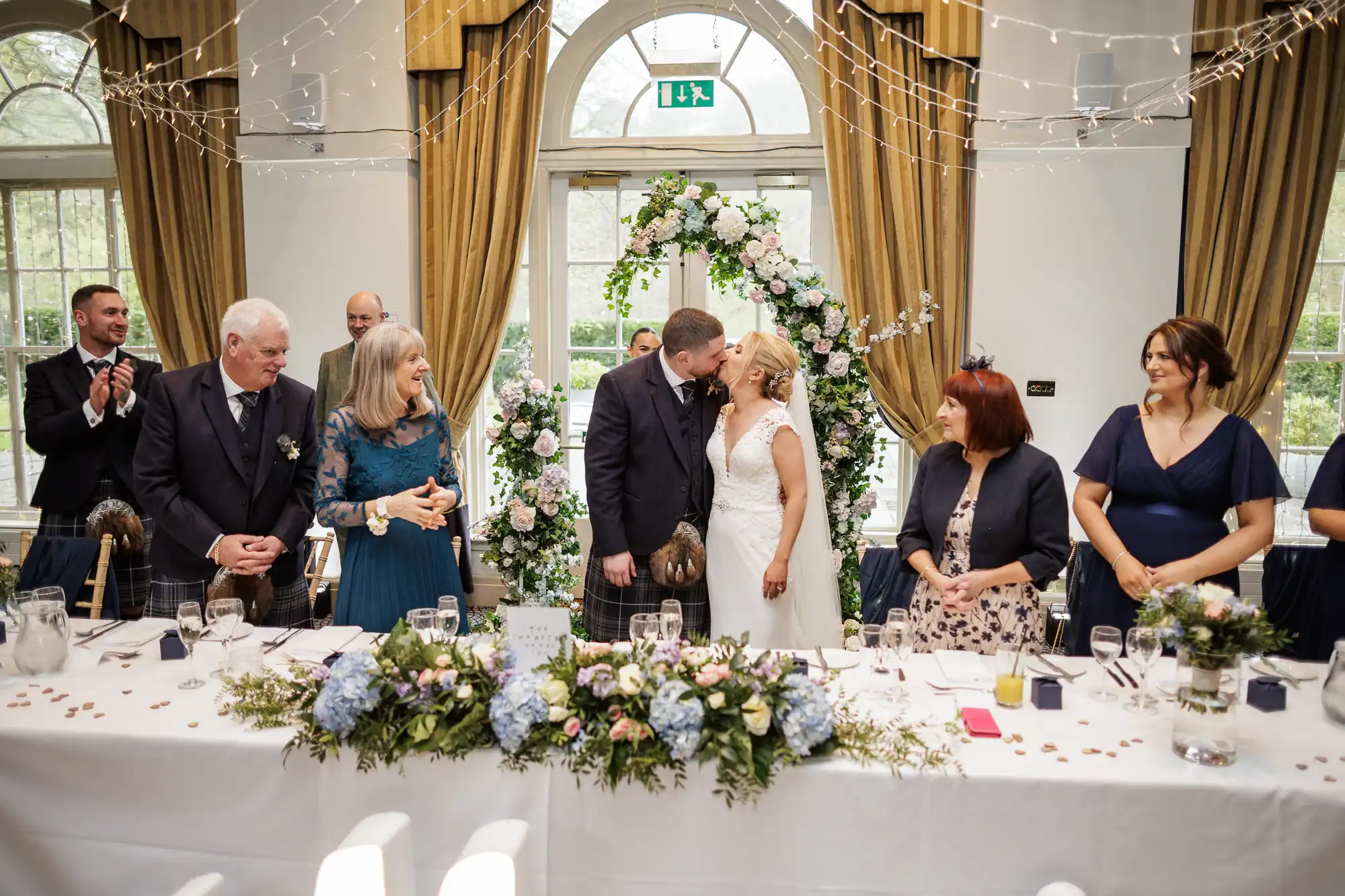 A bride and groom share a kiss at a decorated wedding reception table, surrounded by smiling and applauding guests in formal attire. The room features large windows, gold curtains, fairy lights, and floral arrangements adorning the table and arch behind them.