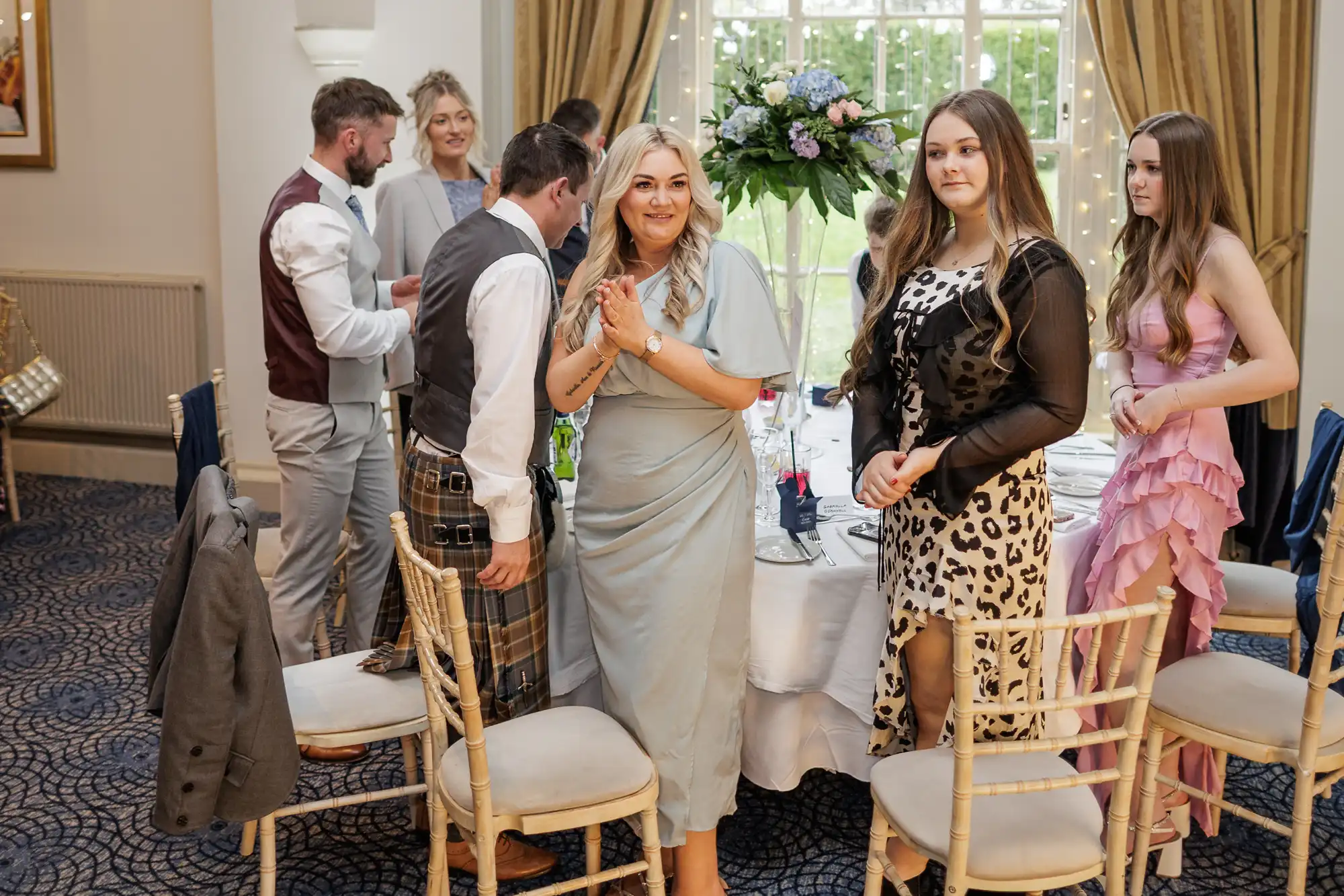A group of people dressed in formal attire stand and smile around a table set for a meal in an elegant, well-lit room with large windows, gold curtains, and a flower centrepiece. The mood is cheerful and social.