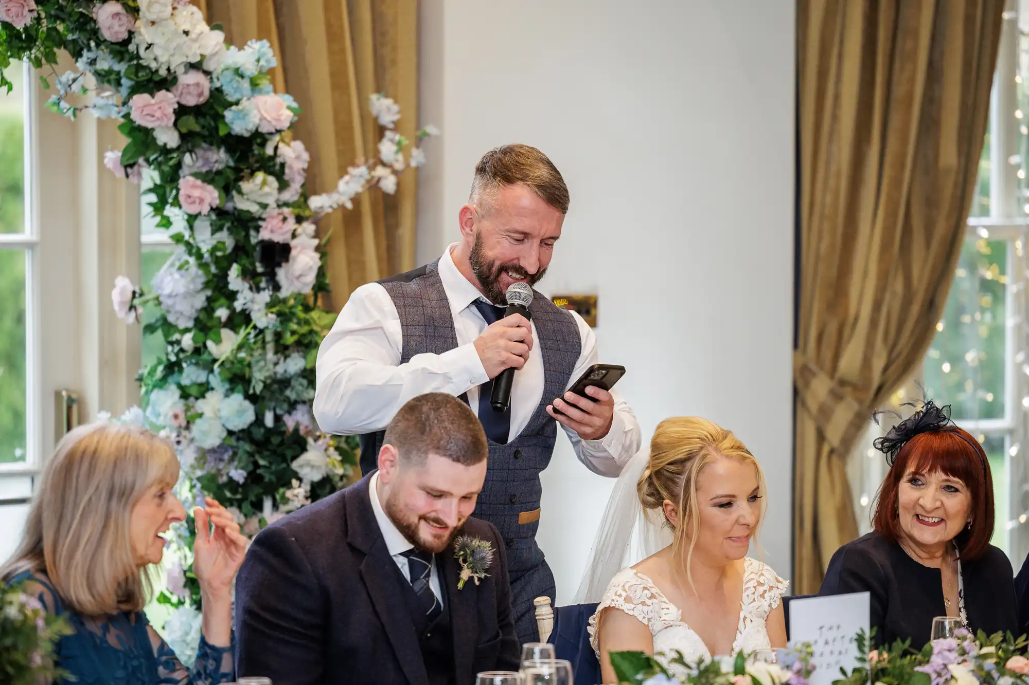 A man in a waistcoat and tie stands holding a microphone and mobile, smiling whilst speaking at a wedding reception table. Around him, guests, including the bride in white and others in formal attire, sit smiling and listening in a flower-decorated room.