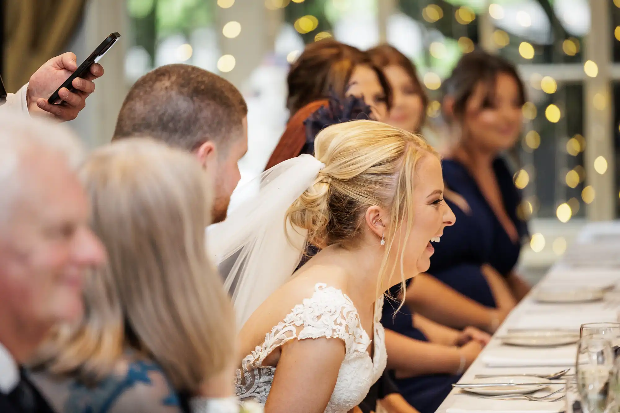 A bride in a white dress and veil laughs joyfully at a decorated reception table, surrounded by guests in formal attire. Soft fairy lights and blurred faces create a warm, celebratory atmosphere.