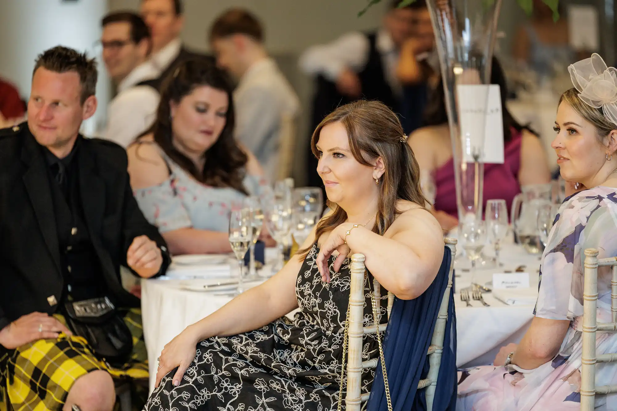 A group of well-dressed guests sit at round tables during a formal event. One woman in a black-patterned dress sits in the foreground, looking attentively ahead. Other guests, including a man in a kilt and women in dresses, are seated around her.