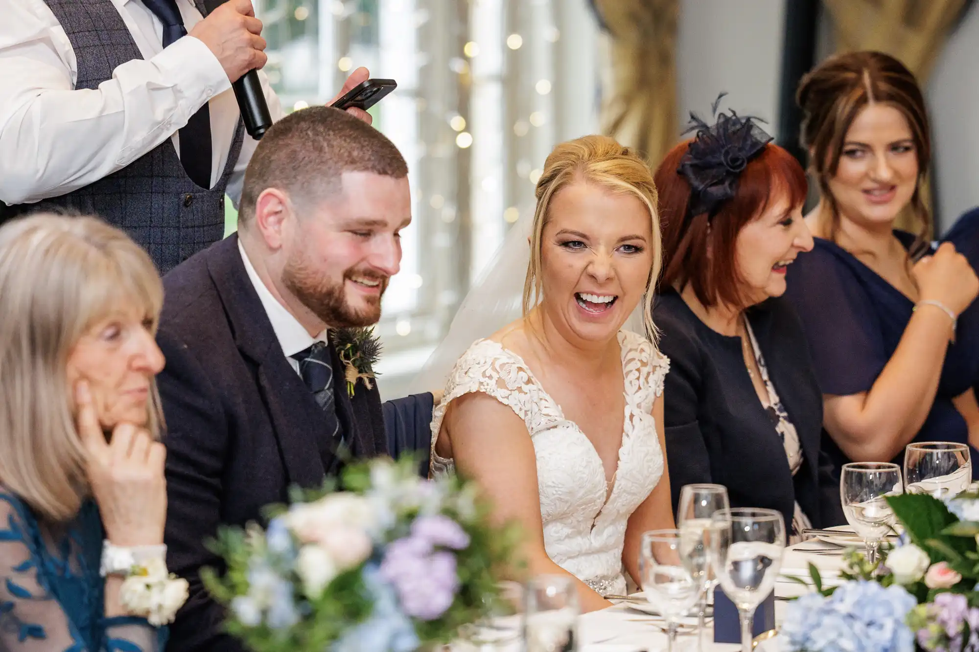 A smiling bride in a white dress sits at a table beside the groom, surrounded by guests in formal attire. They laugh together at a decorated reception with flowers, wine glasses, and a man speaking into a microphone in the background.