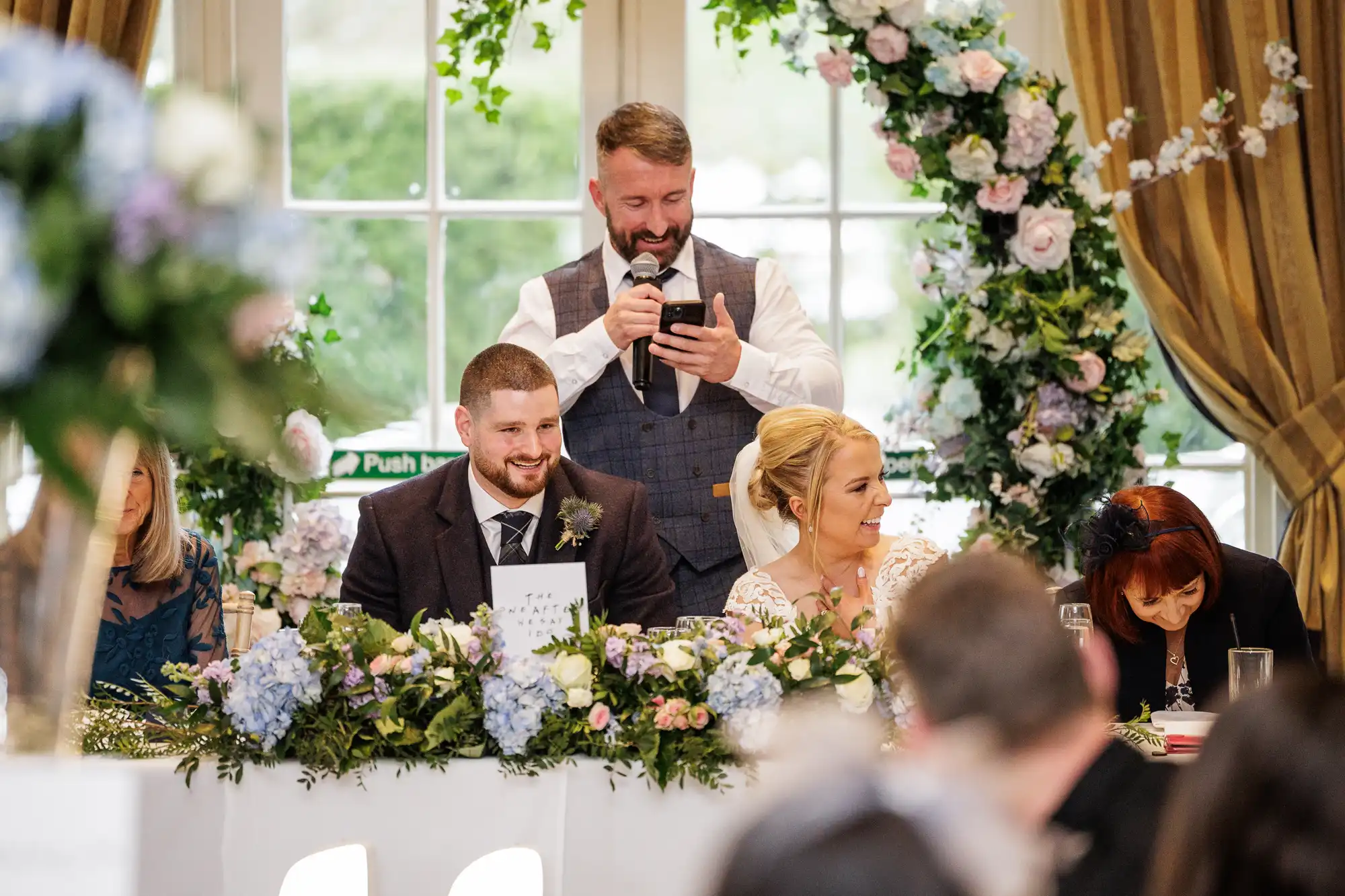A man stands behind a wedding top table, smiling as he reads from his mobile into a microphone. The bride and groom sit in front, surrounded by floral arrangements, laughing with guests. An arch of flowers and large windows are in the background.