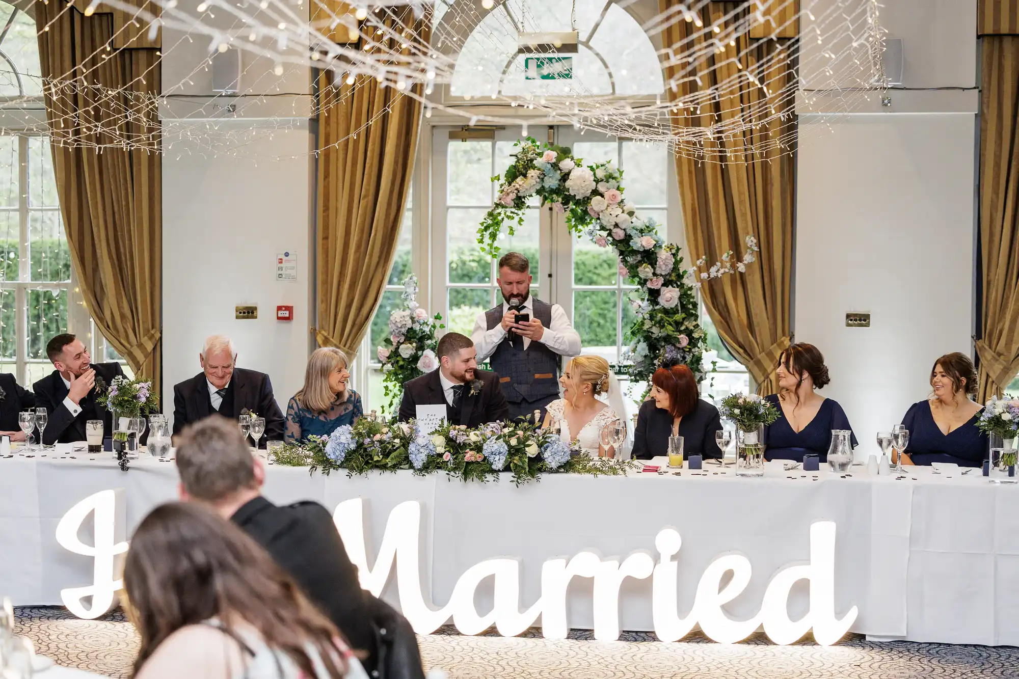 A wedding reception top table with eight people dressed formally, seated in front of a large illuminated "Just Married" sign. A man stands behind, taking a photo. Floral decorations and draped curtains frame the festive, elegant indoor scene.
