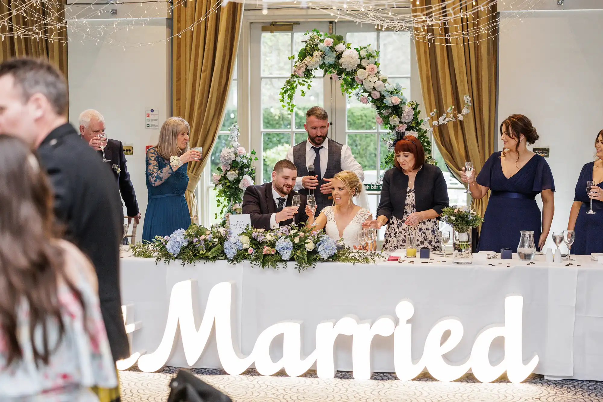 A bride and groom sit at a flower-adorned table, surrounded by family and friends raising glasses in a toast. Large illuminated “Married” letters are in front. Gold curtains frame tall windows and a floral arch stands behind the couple, creating a joyful wedding scene.