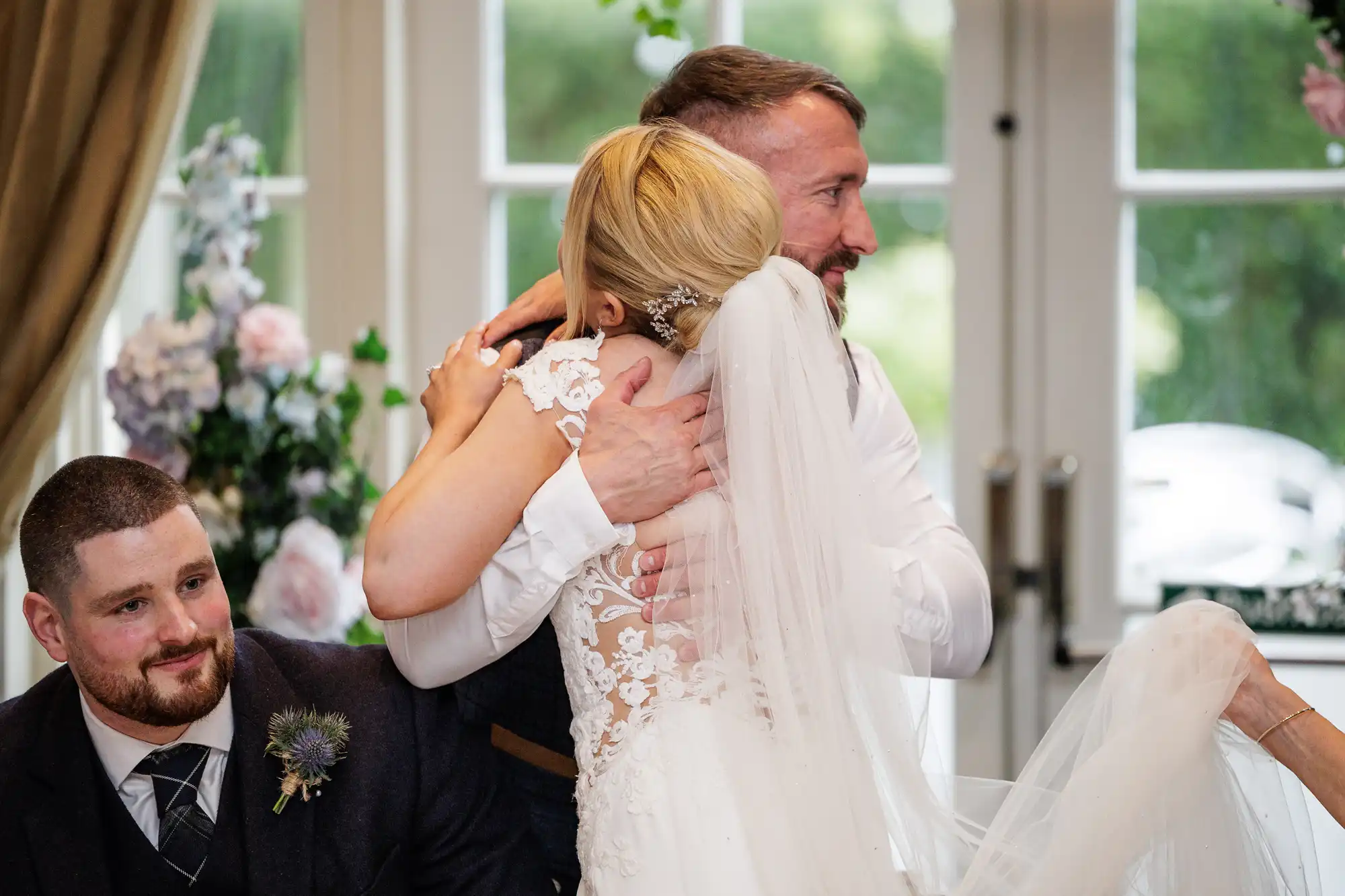 A bride in a white lace wedding dress hugs a man in a waistcoat, her back to the camera. A man with a beard and short hair sits nearby, smiling. They are indoors, with floral decorations and large windows in the background.