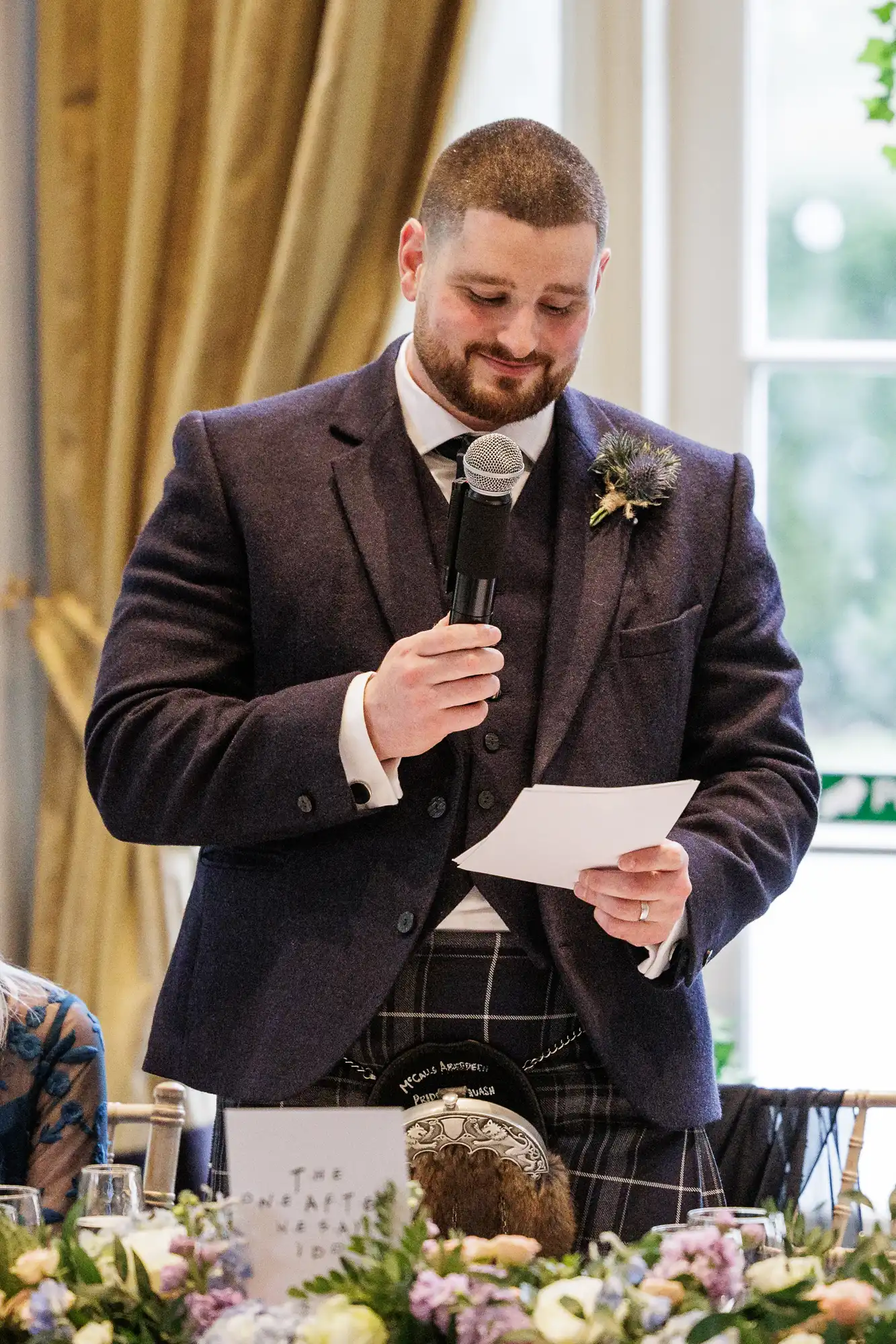 A bearded man in a kilt and dark jacket holds a microphone and reads from a paper, smiling. He stands at a decorated table with flowers and glassware, in a room with gold curtains and large windows behind him.