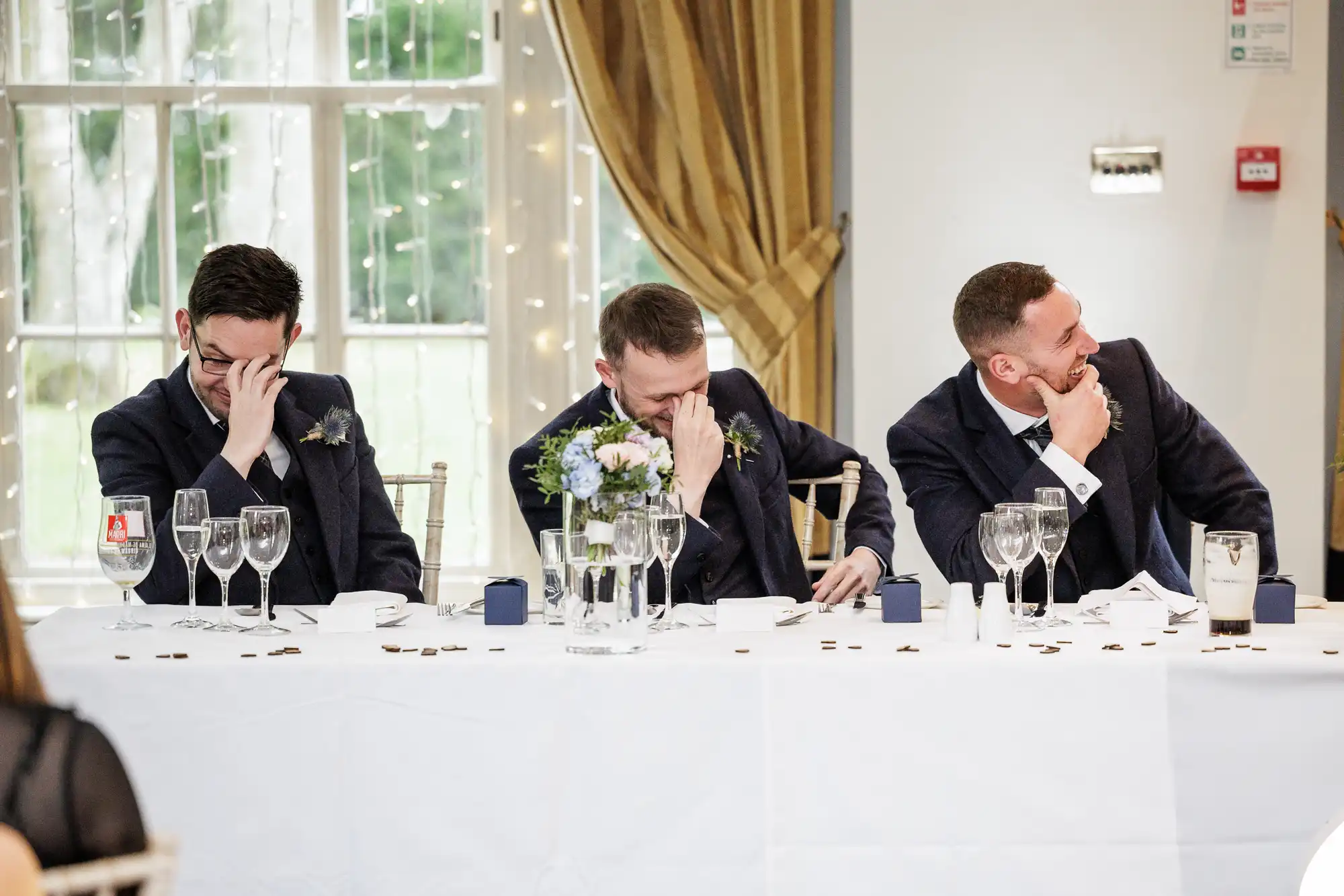 Three men in dark suits sit at a wedding top table, laughing and covering their faces with their hands. The table is set with glasses, flowers, and decorations, with a large window and gold curtains in the background. The mood appears joyful and light-hearted.